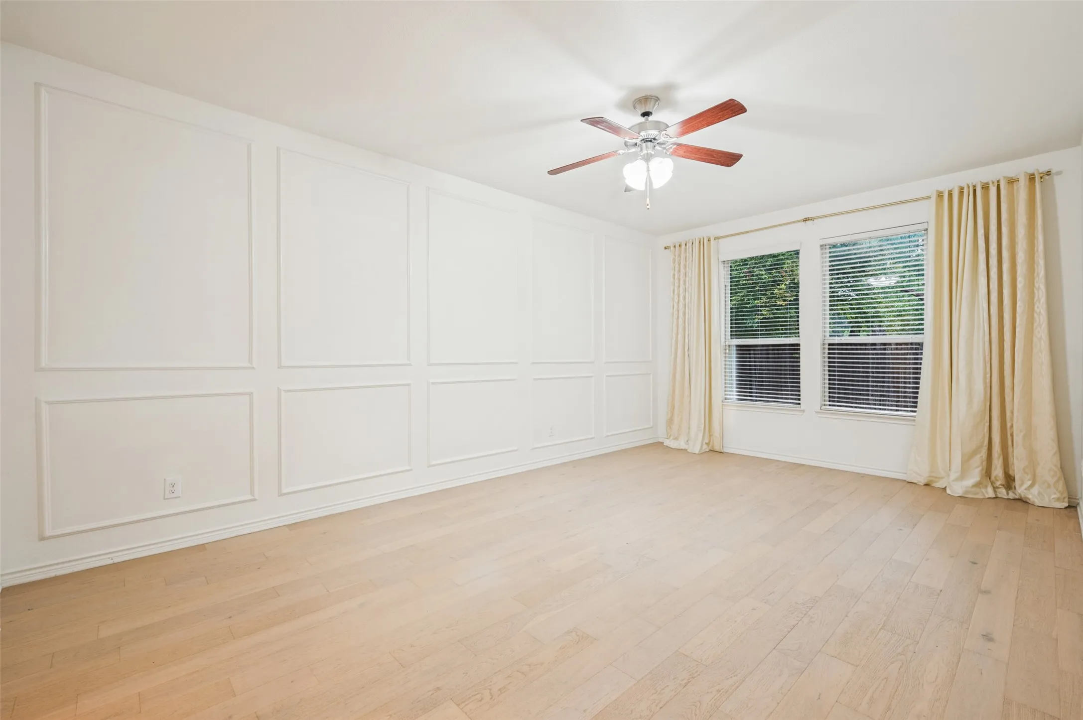 Light wood-finish flooring, accent wall with decorative molding, two windows with blinds, and a ceiling fan with light fixture