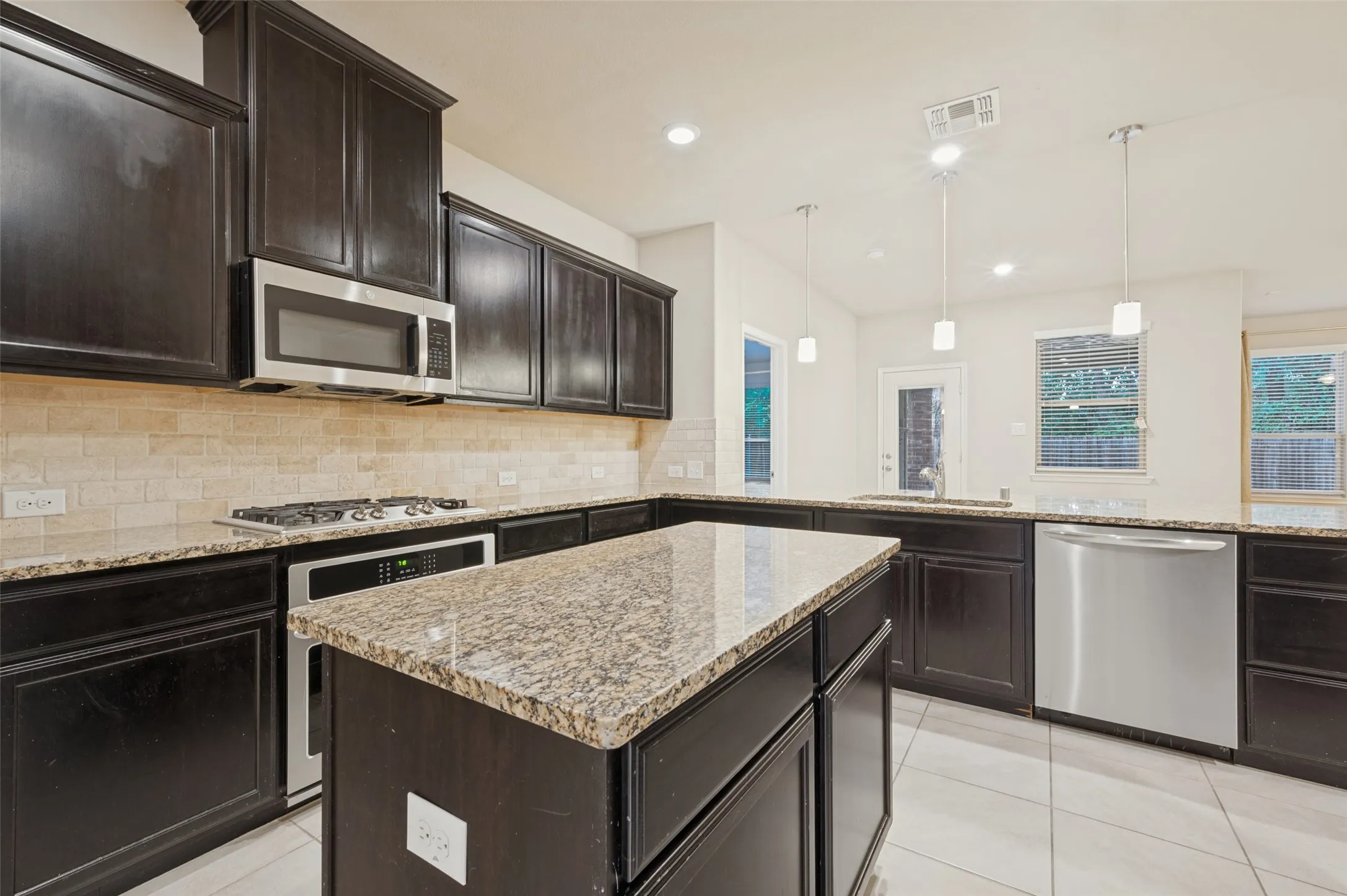 Kitchen featuring dark wood cabinetry, granite countertops, tile backsplash, and stainless steel appliances including a microwave, gas range, and dishwasher