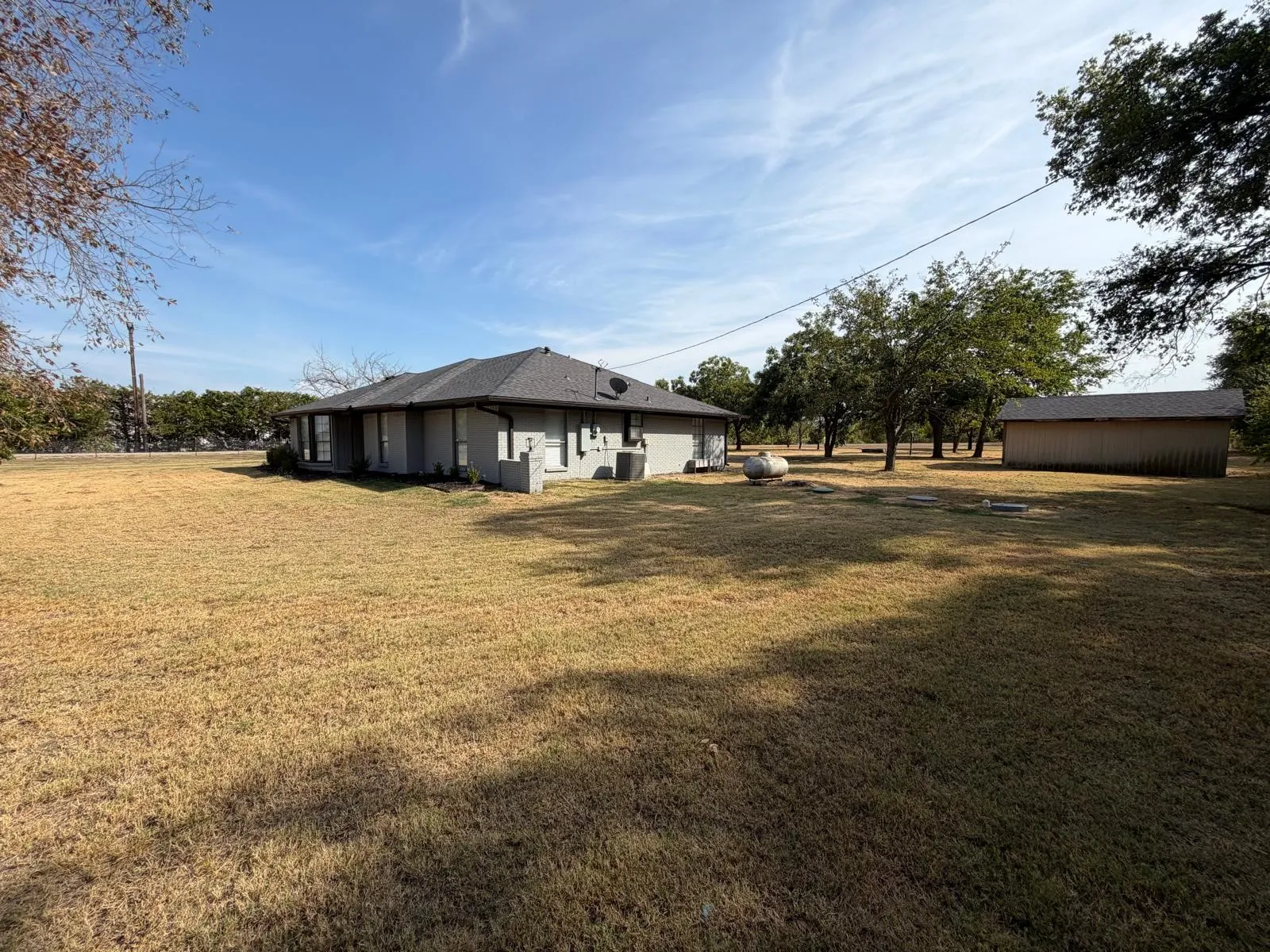Single-story residence with a gray shingle roof, light-colored exterior siding, multiple windows, and a detached outbuilding