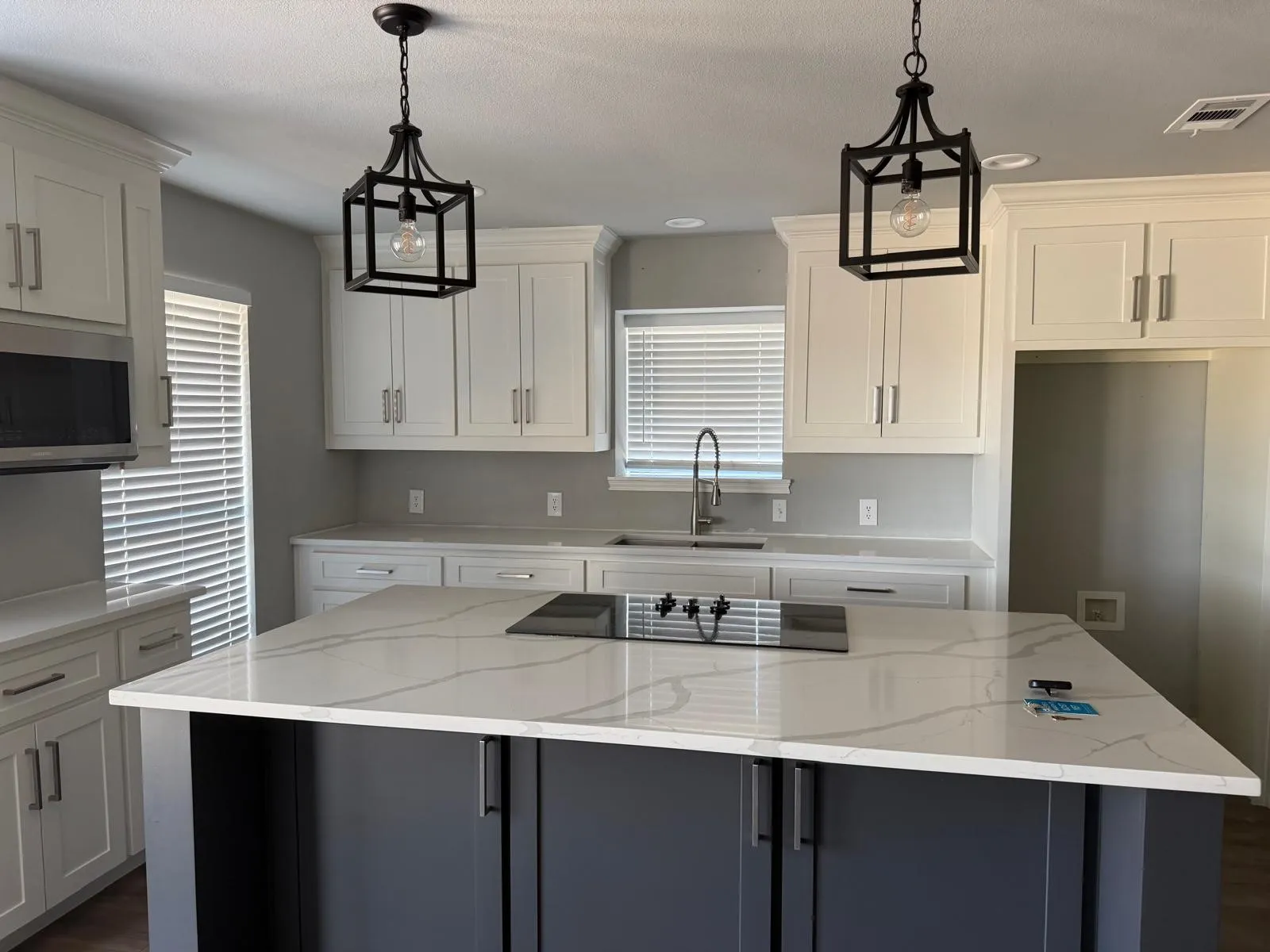 Kitchen featuring white shaker cabinetry, an island with a built-in cooktop, light-colored countertops with veining, a stainless steel microwave, and pendant lighting
