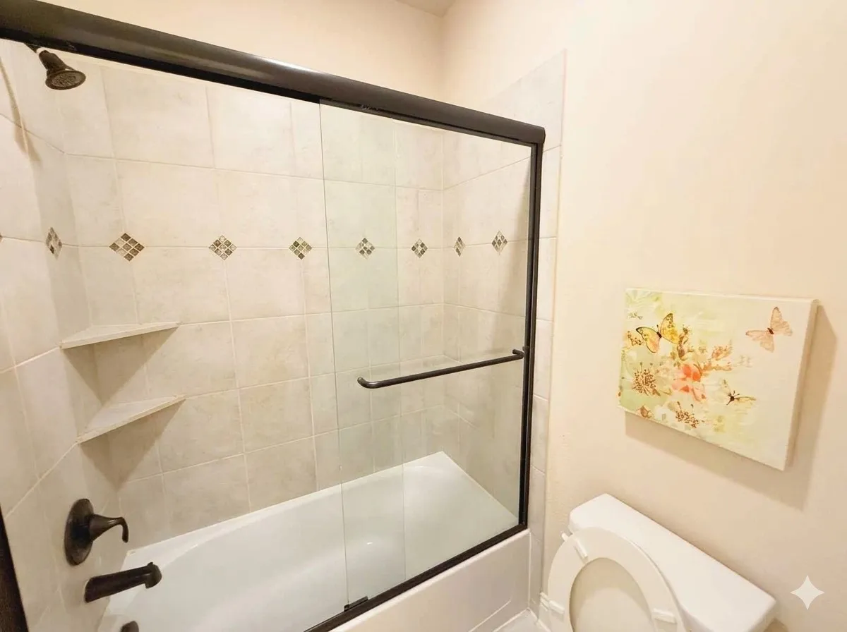 Bathtub with sliding glass shower door, tiled surround with decorative accents, oil-rubbed bronze fixtures, and two built-in corner shelves