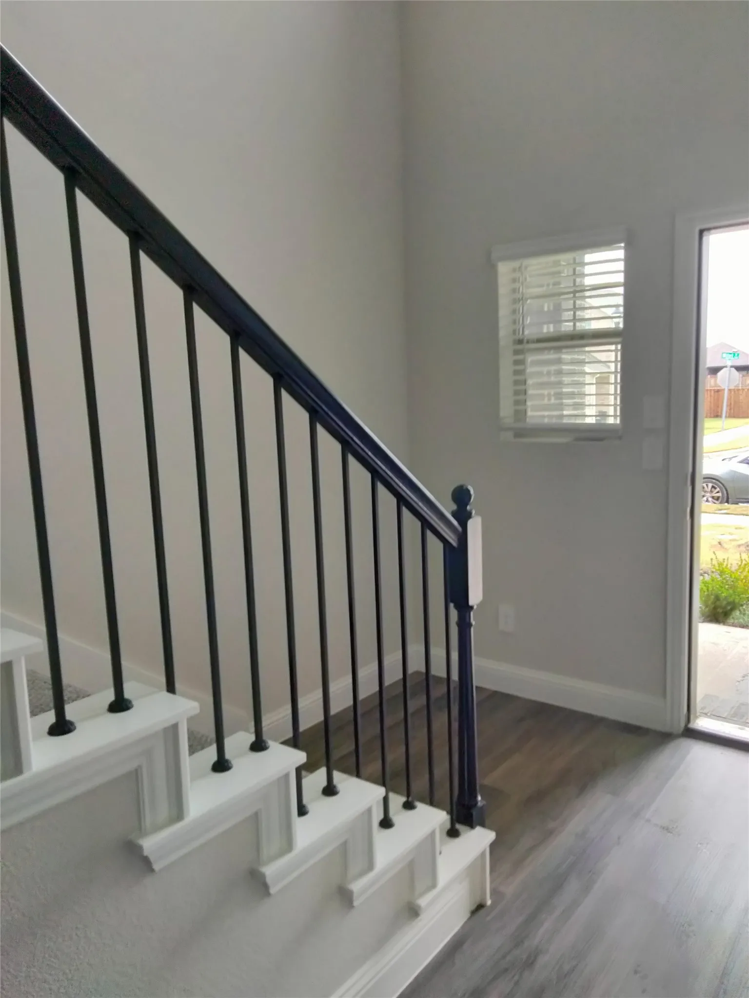 Staircase with black metal balusters and white risers, carpeted treads, wood-finish flooring, and white window blinds