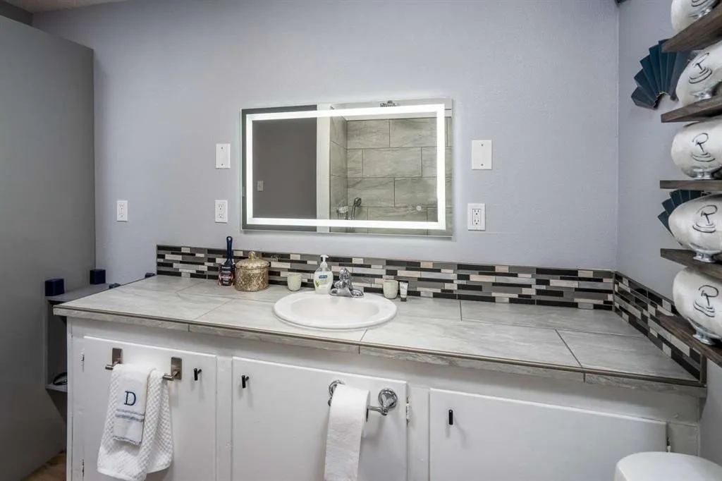 Illuminated vanity mirror, single basin sink, tiled countertop, mosaic tile backsplash, and white cabinetry