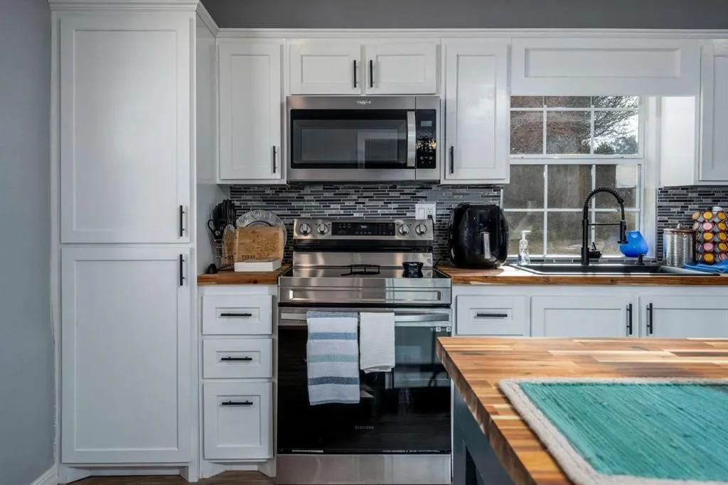 White shaker-style cabinetry with matte black hardware, stainless steel appliances, mosaic tile backsplash, wood-finish countertops, and an undermount sink with a matte black faucet
