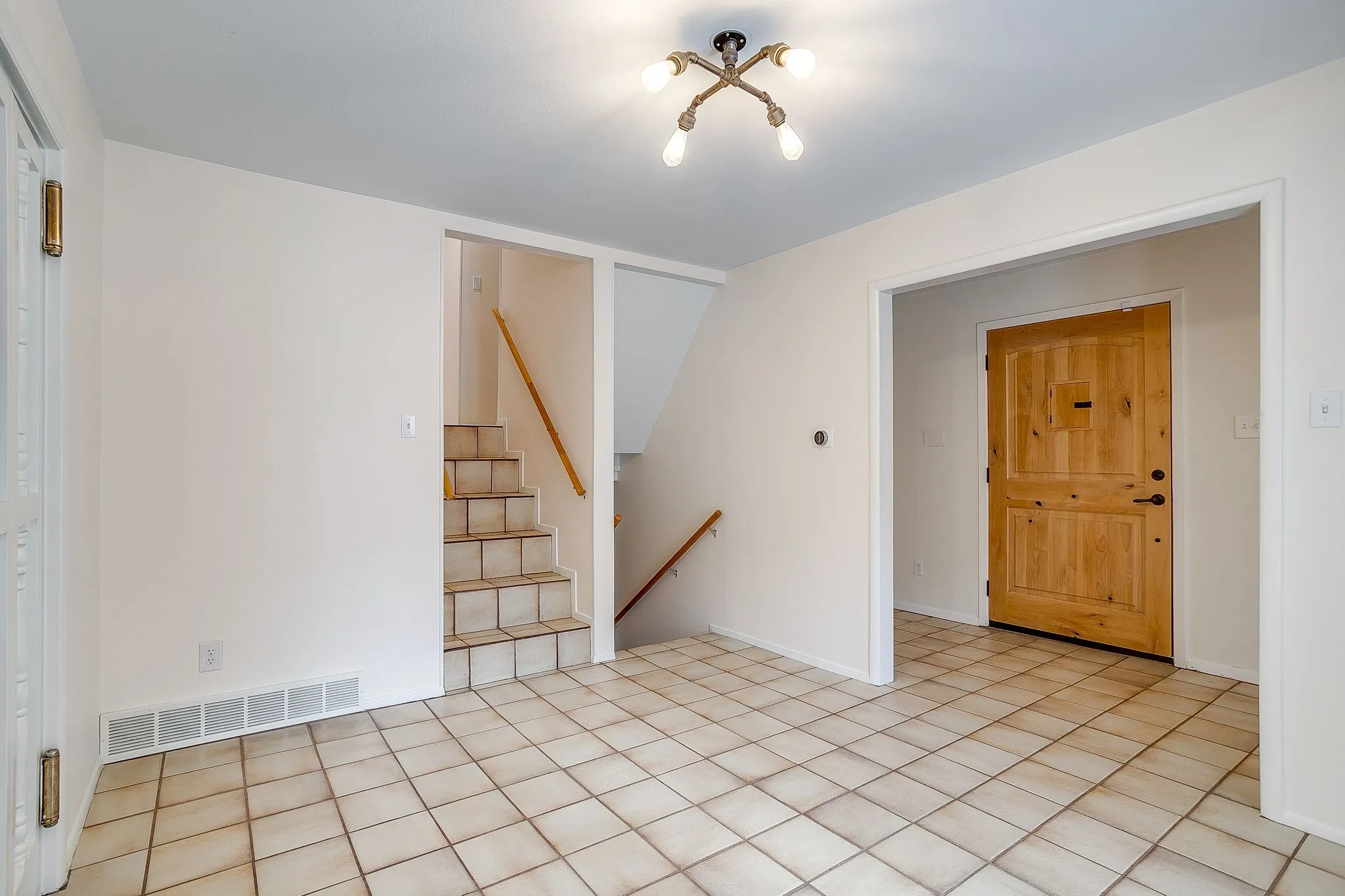 Tiled flooring, light wood-tone entry door, staircase with wood-tone railing, and a multi-bulb ceiling fixture