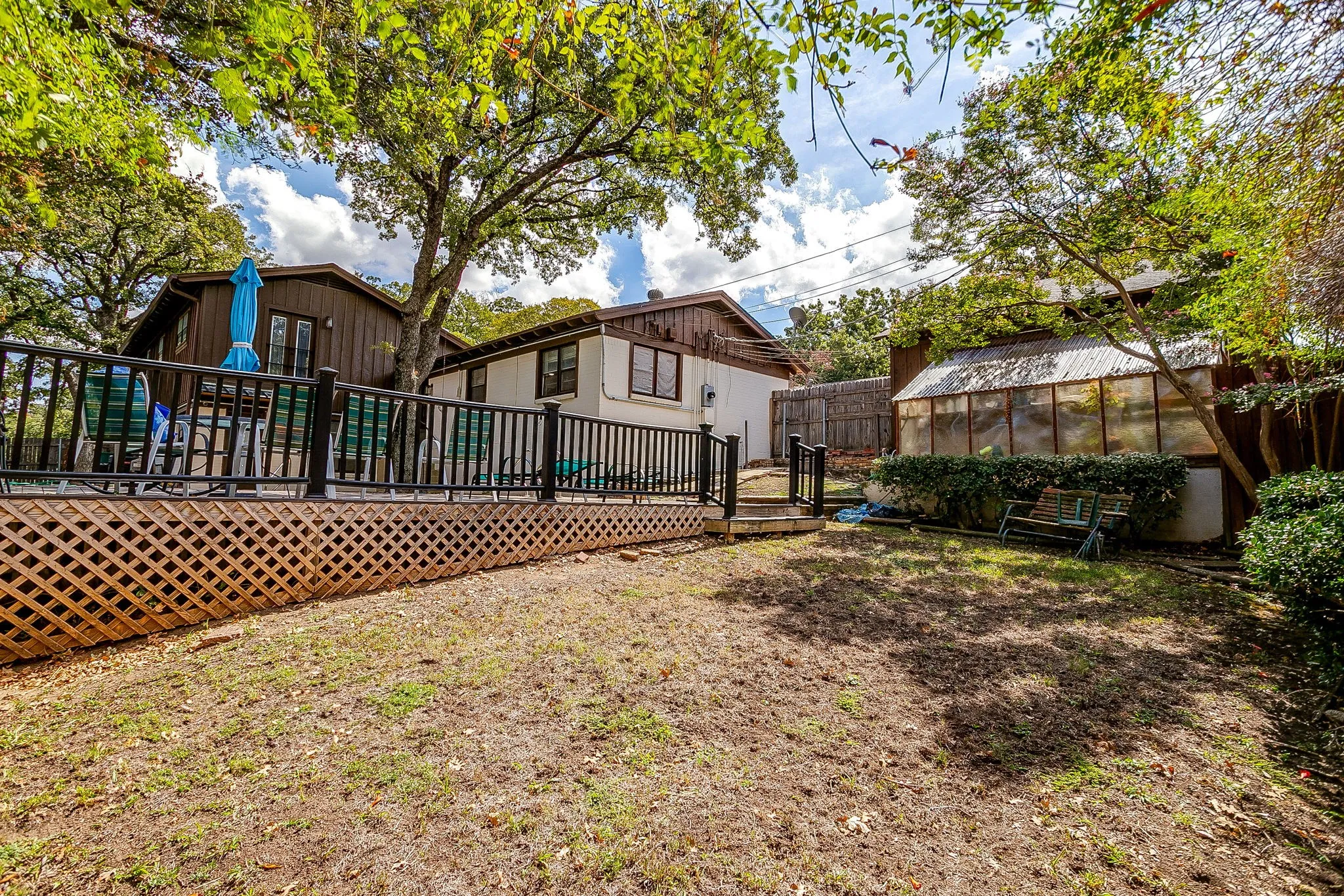 Elevated wood deck with lattice skirting and black railings, leading to a spacious yard with a greenhouse structure and mature trees