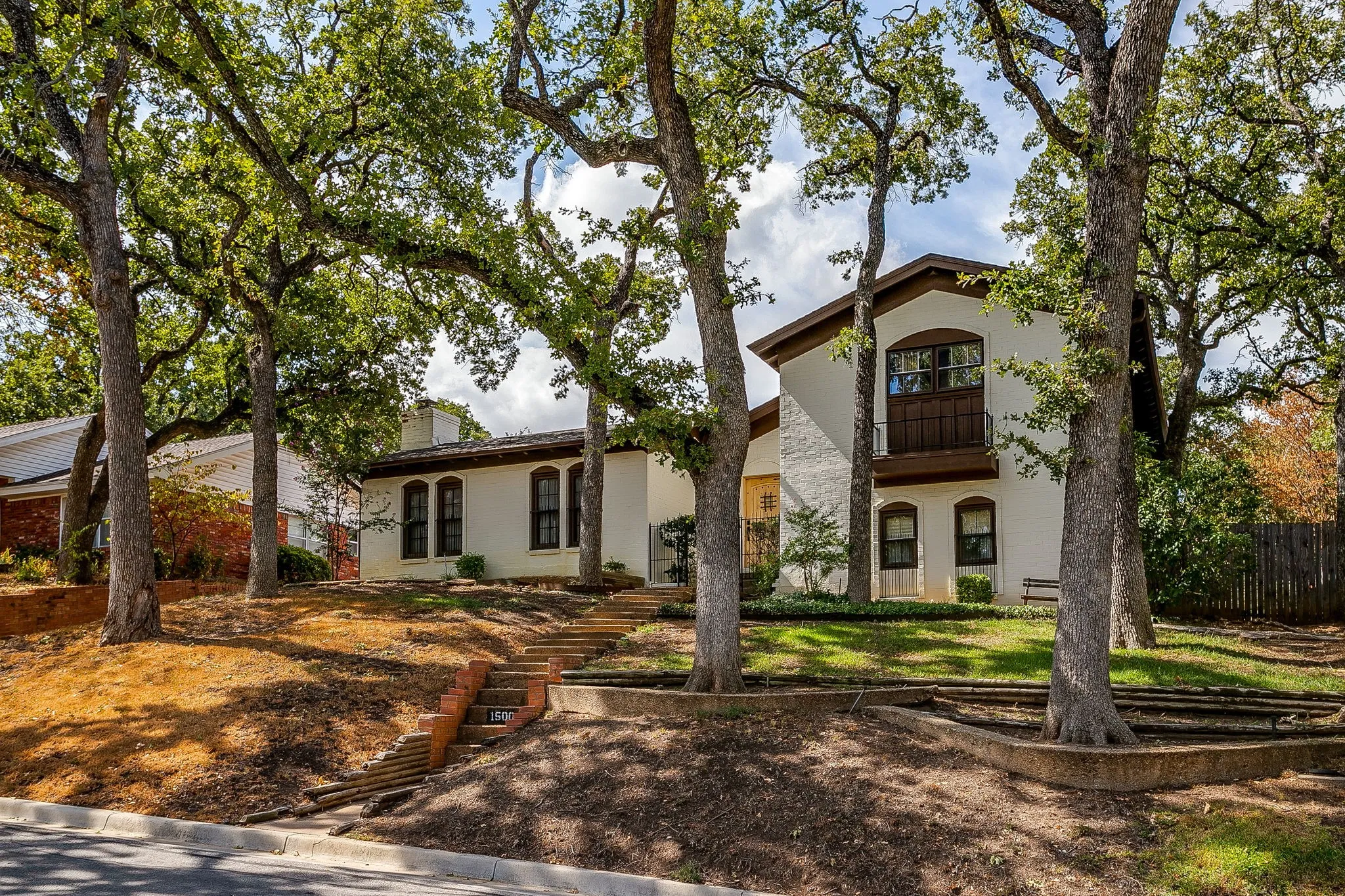 White stucco exterior, brown trim accents, arched window casings, wood-finish balcony, tiered landscaping with mature trees