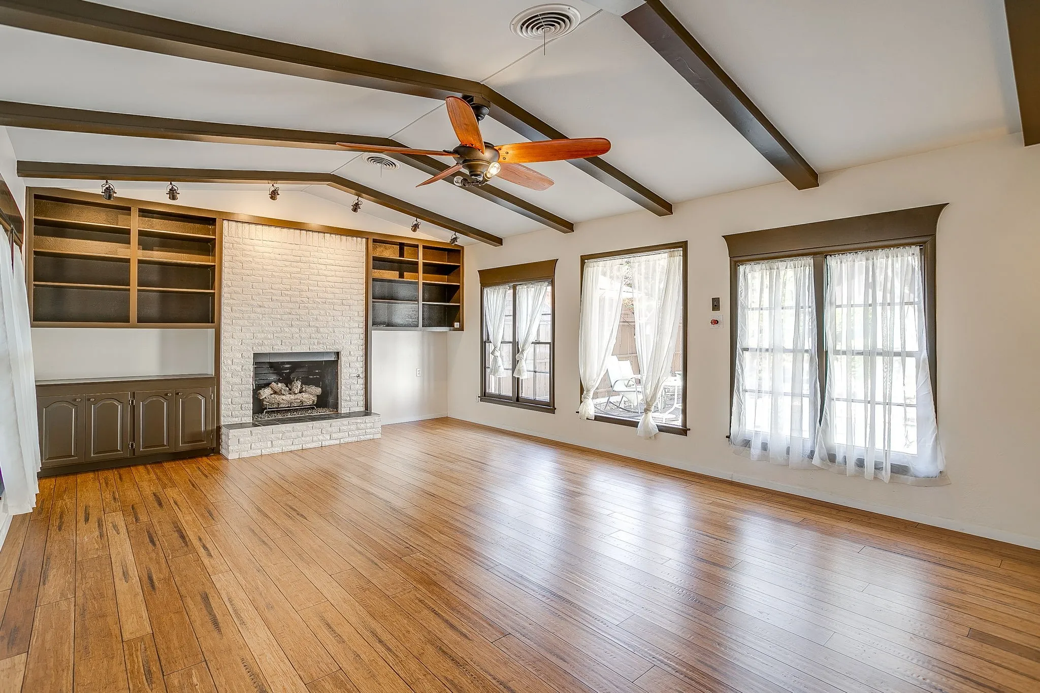 Vaulted ceiling with exposed beams, wood-finish flooring, white brick fireplace with hearth, built-in shelving, and track lighting