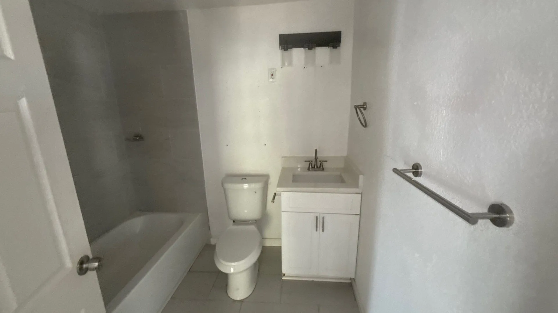 White vanity with a single basin sink, wall-mounted light fixture, white toilet, and tub with tiled surround