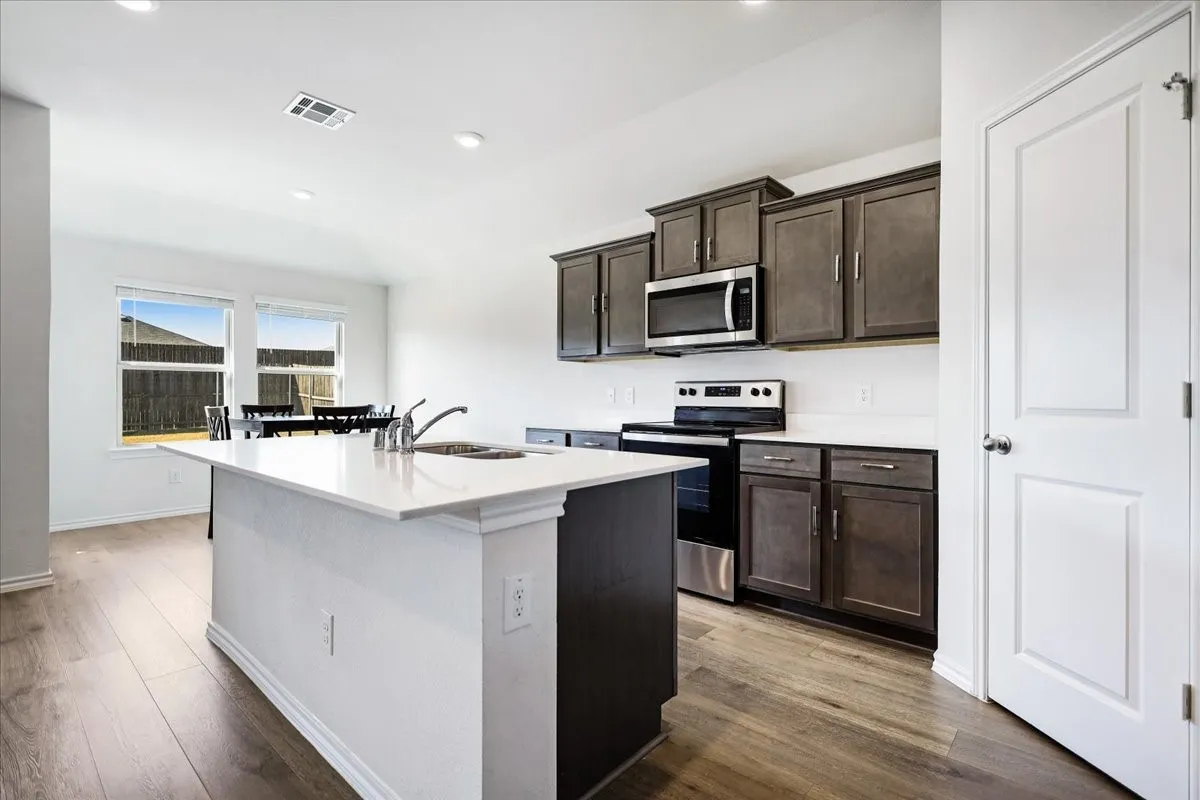 Kitchen featuring a central island with an integrated sink, wood-finish flooring, white countertops, built-in stainless steel microwave, and dark-toned cabinetry
