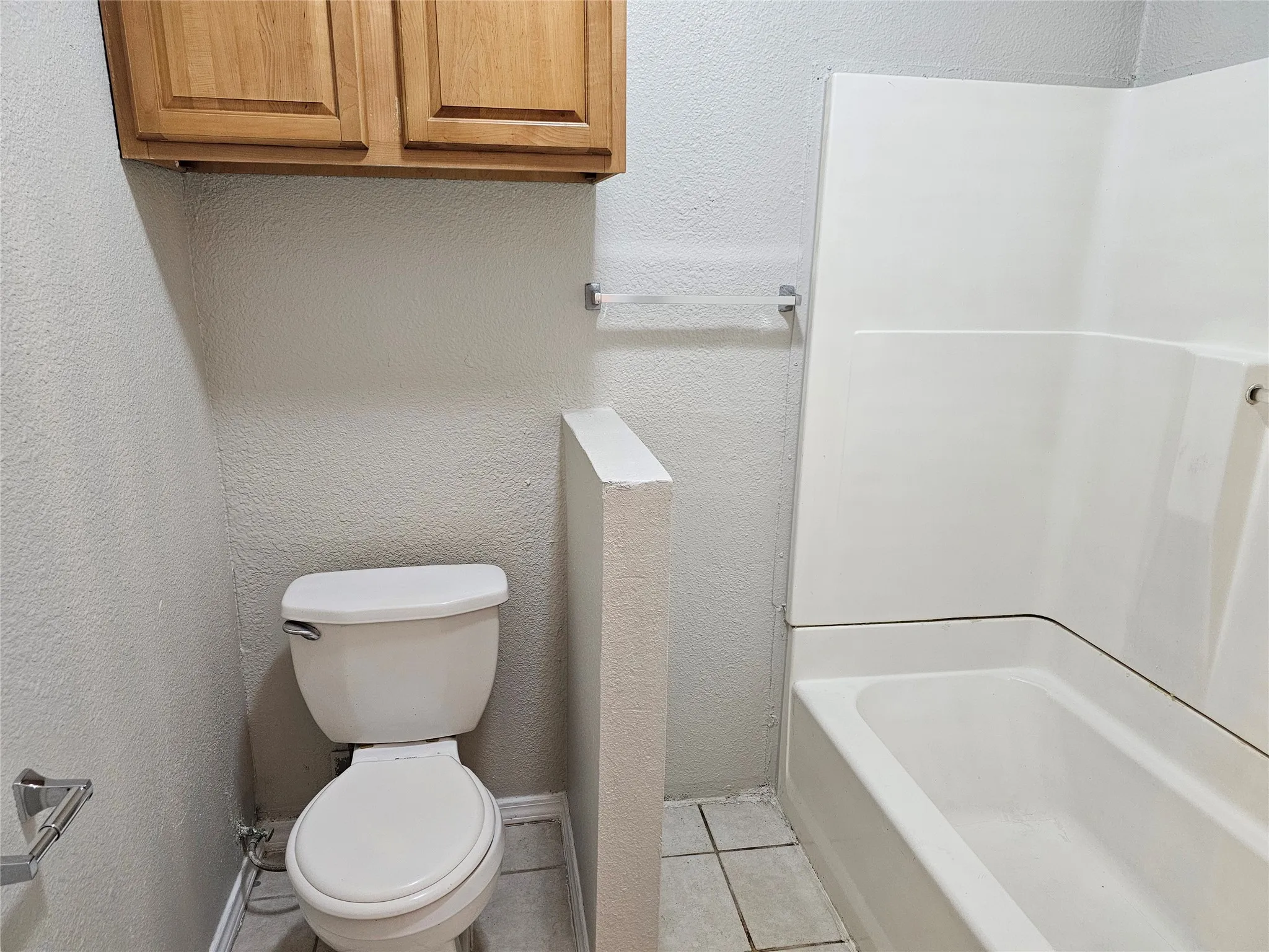 White ceramic toilet, integrated white bathtub and shower surround, light wood-finish cabinetry, and tiled flooring
