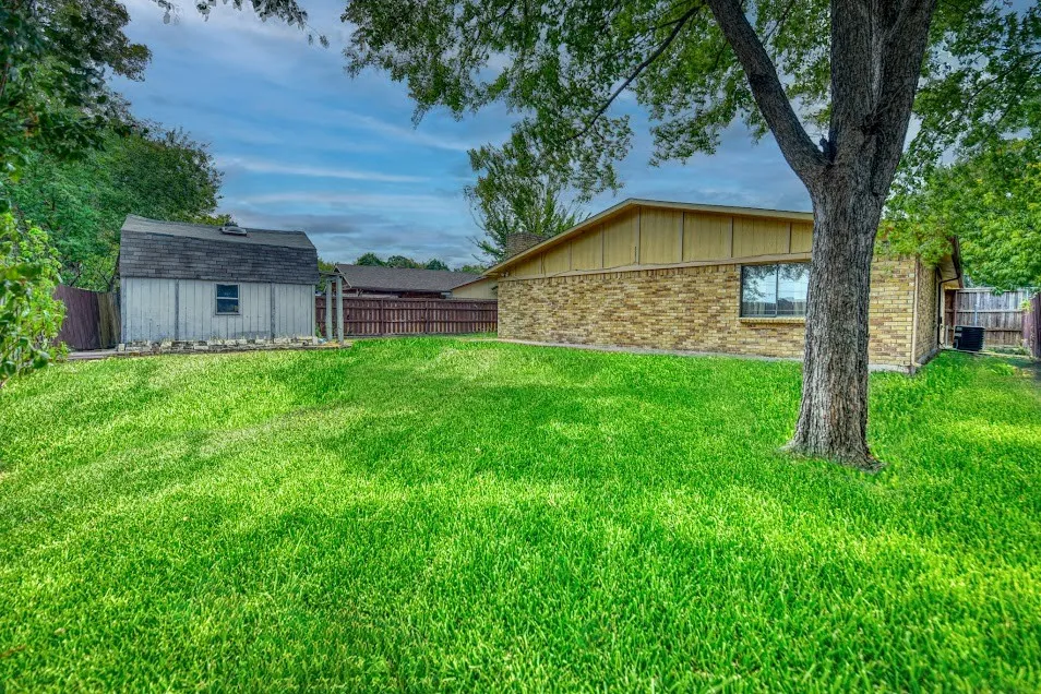 Expansive lawn with a mature shade tree, featuring a detached storage shed and a brick exterior home