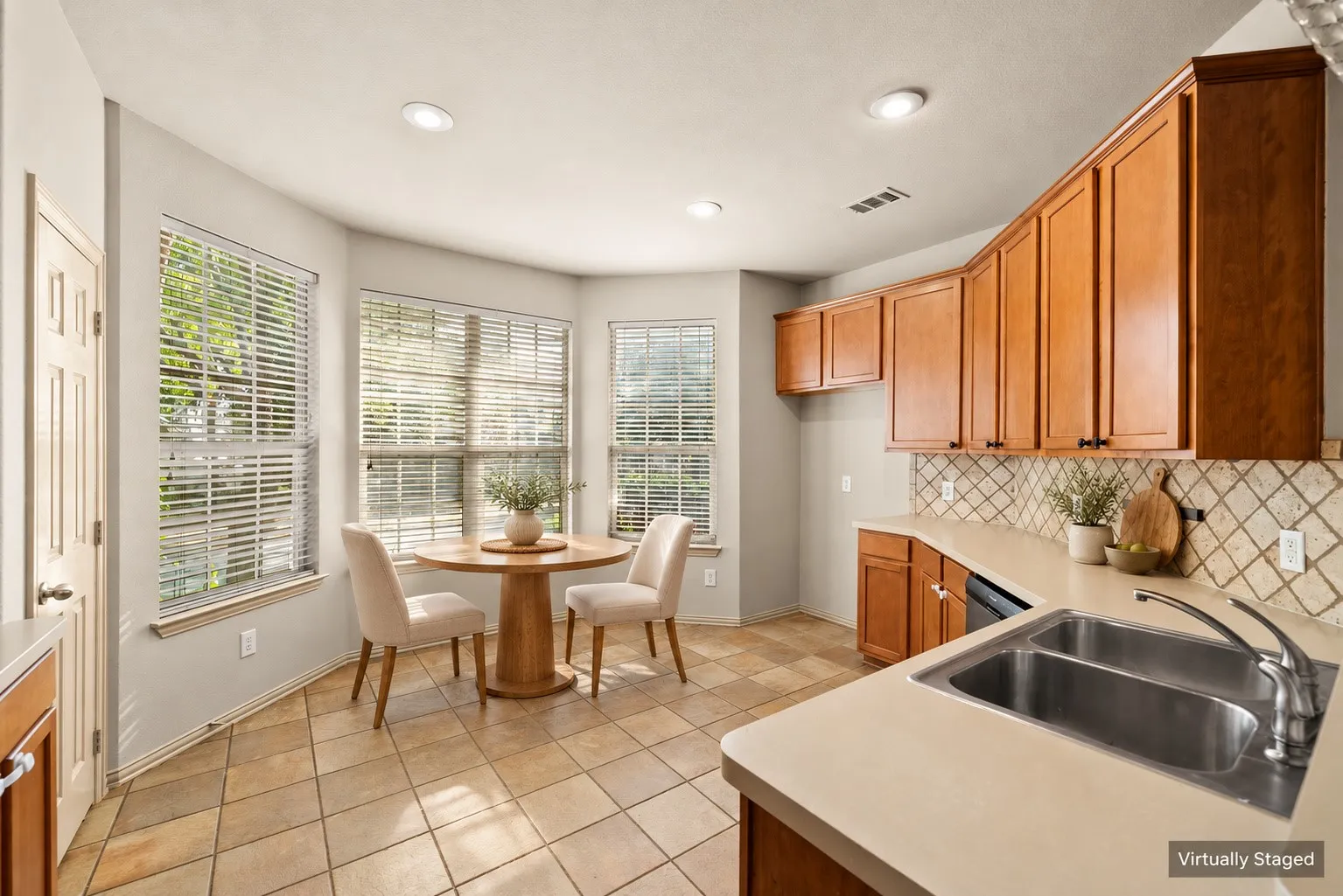 Light wood-finish cabinetry with dark hardware, dual basin stainless steel sink, light-toned countertops, tiled backsplash, and recessed lighting