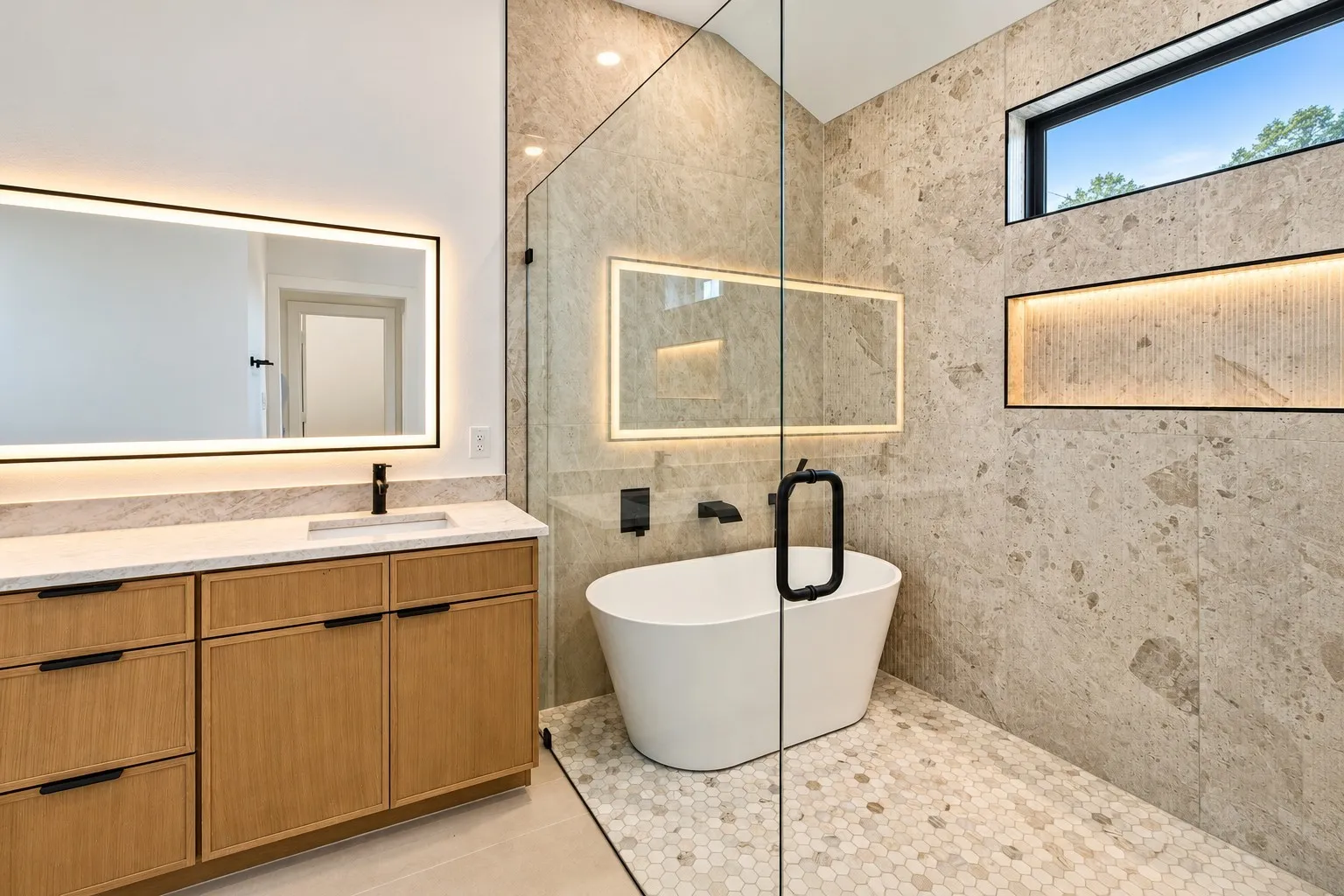 Freestanding soaking tub within a glass-enclosed shower, complemented by large format wall tile and hexagon mosaic shower flooring