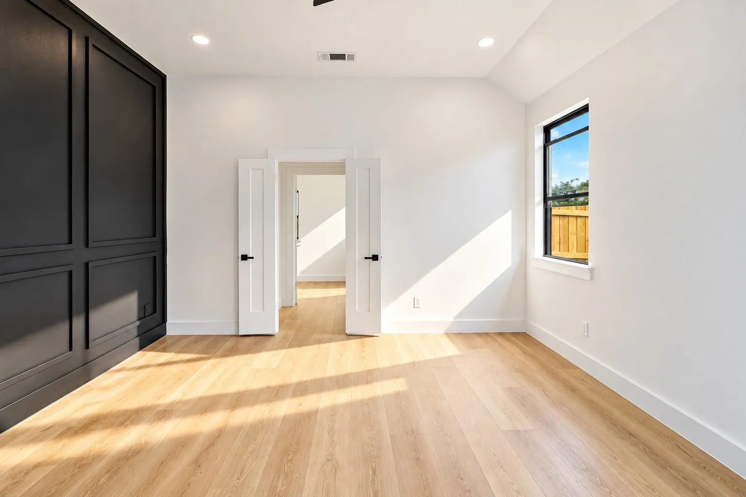 Wood-finish flooring, recessed lighting, accent wall with panel molding, black-framed window with wood fence backdrop, and double doors with black hardware