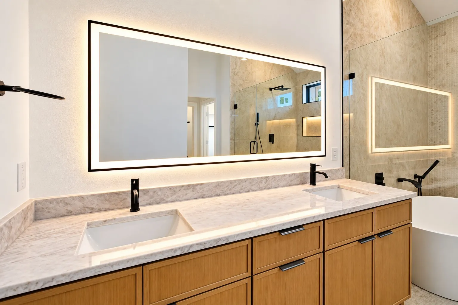 Dual vanity with integrated sinks, light-toned stone countertop, wood-finish cabinetry with dark hardware, back-lit mirror, and matte black fixtures