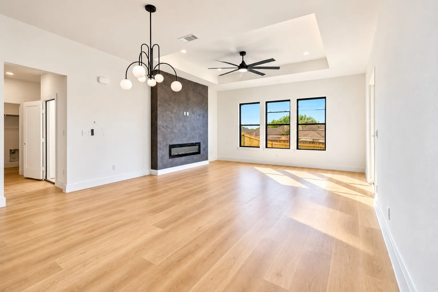 Expansive living area featuring wood-finish flooring, a contemporary chandelier, a ceiling fan, an electric fireplace with a textured surround, and three large windows