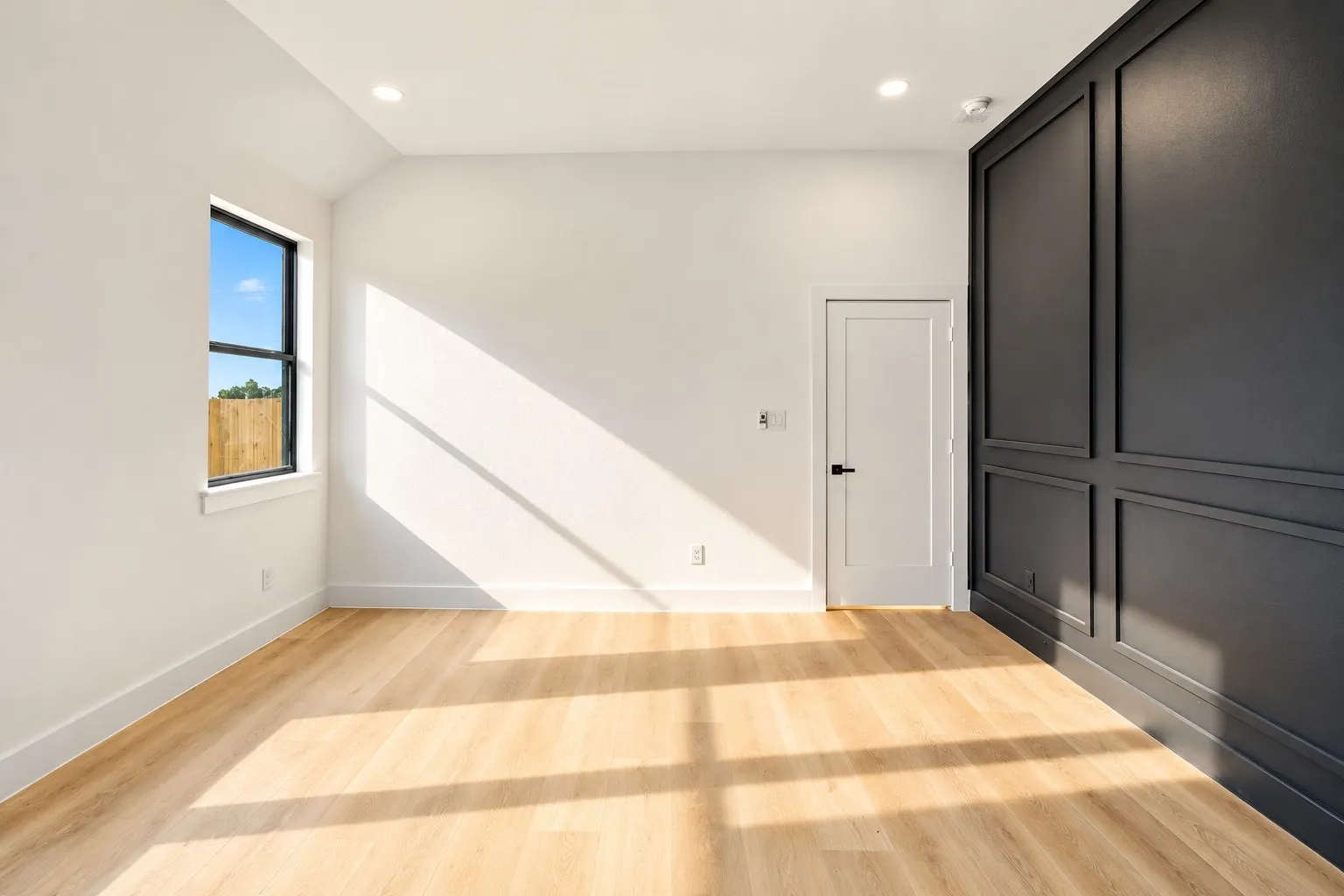 Wood-finish flooring, recessed lighting, accent wall with panel molding, single window with black trim, and a white flat-panel door with black hardware