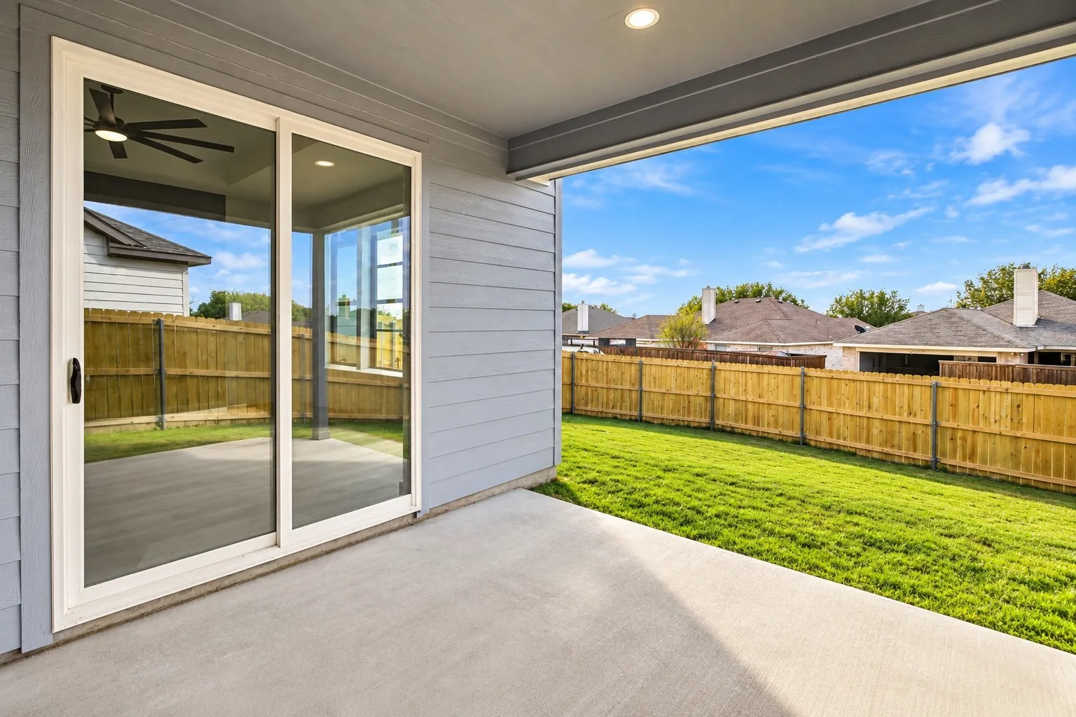 Covered patio with a concrete surface, sliding glass doors, and a wood fence enclosing a grass lawn
