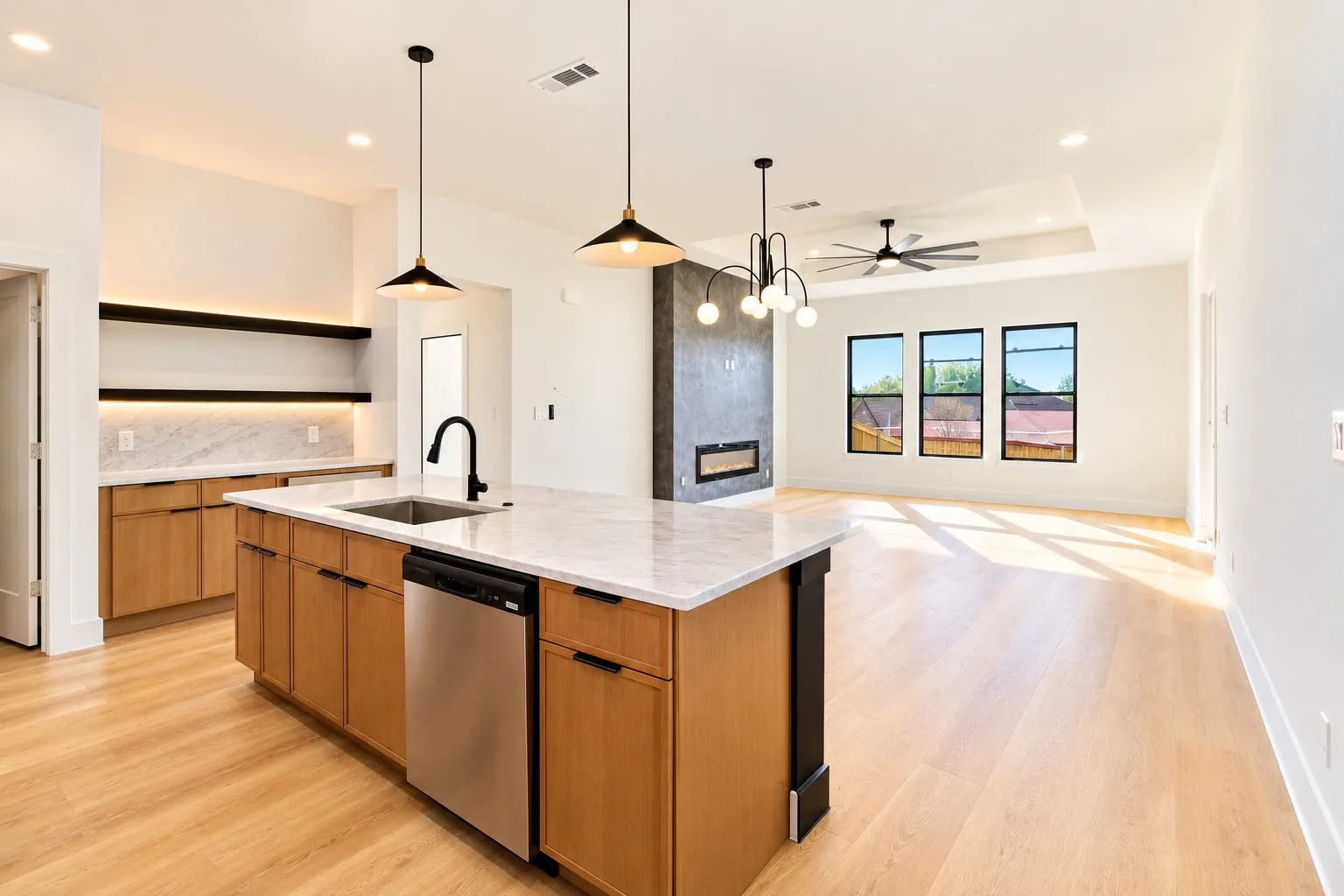 Open-concept interior featuring light wood-finish flooring, a kitchen island with a built-in sink and stainless steel dishwasher, a built-in fireplace with a dark surround, and multiple windows