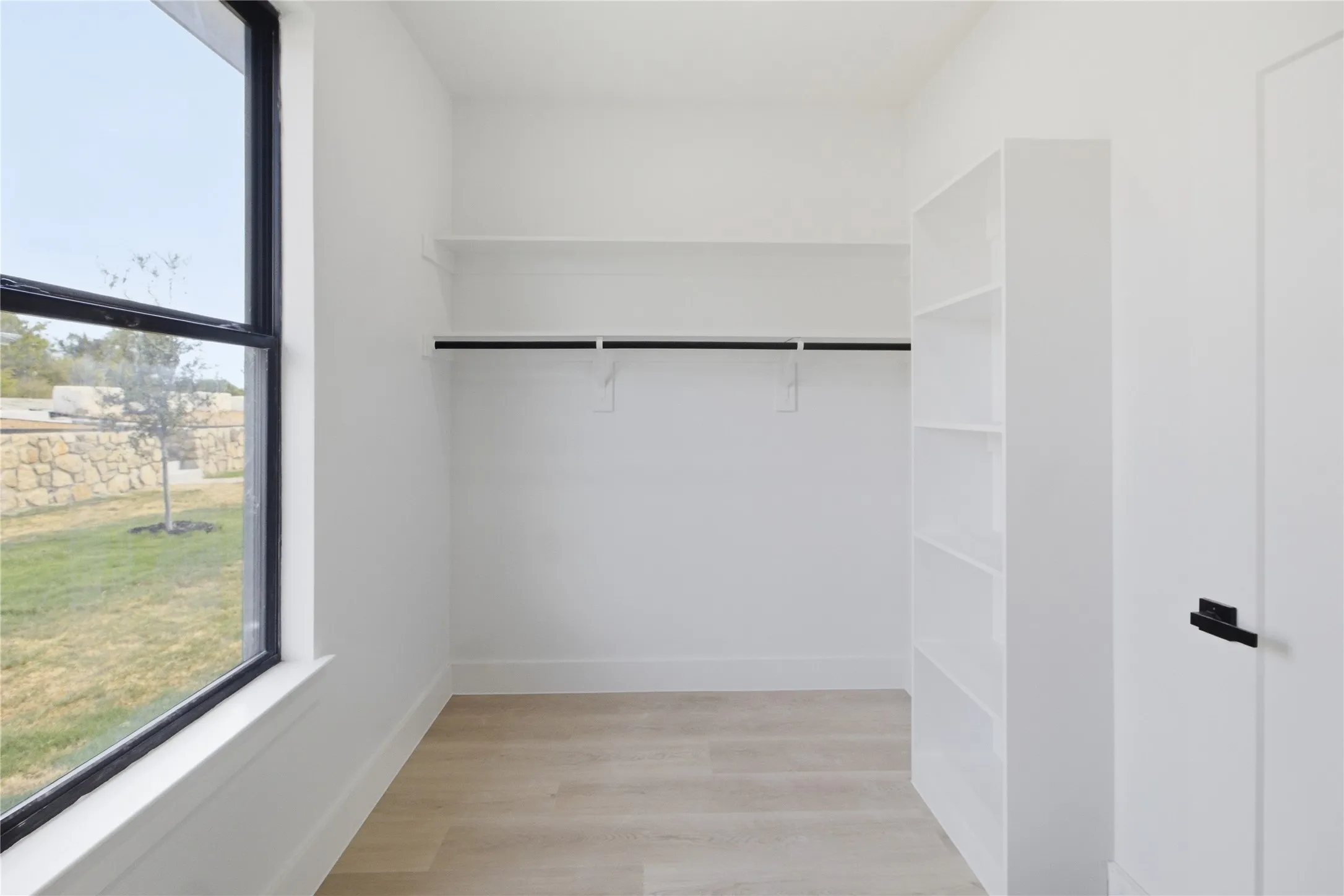 Light wood-finish flooring, built-in shelving, white walls, and a large window with black framing