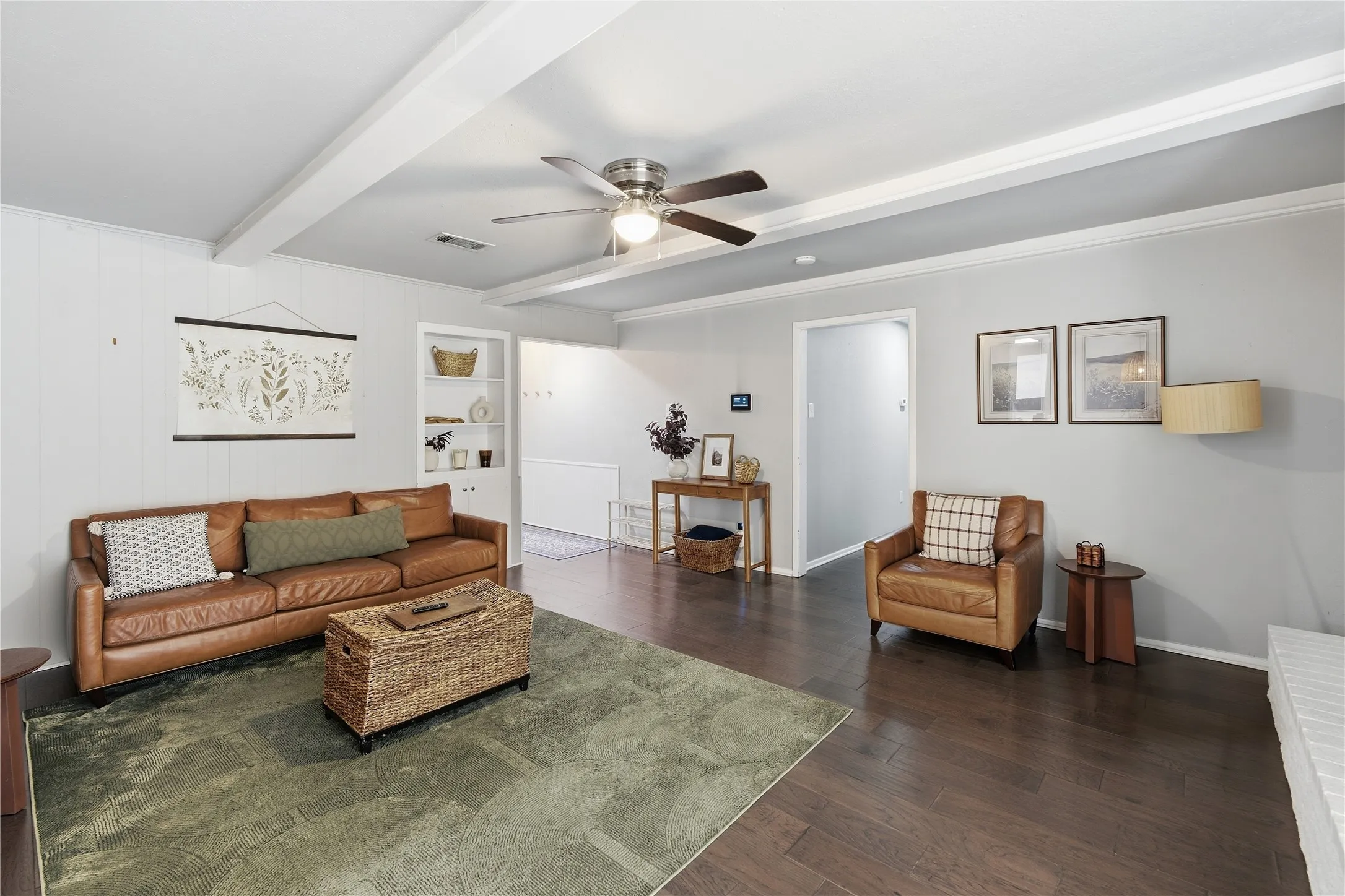 Dark wood-finish flooring, white exposed ceiling beams, built-in shelving, white panel accent wall, and a ceiling fan with integrated lighting