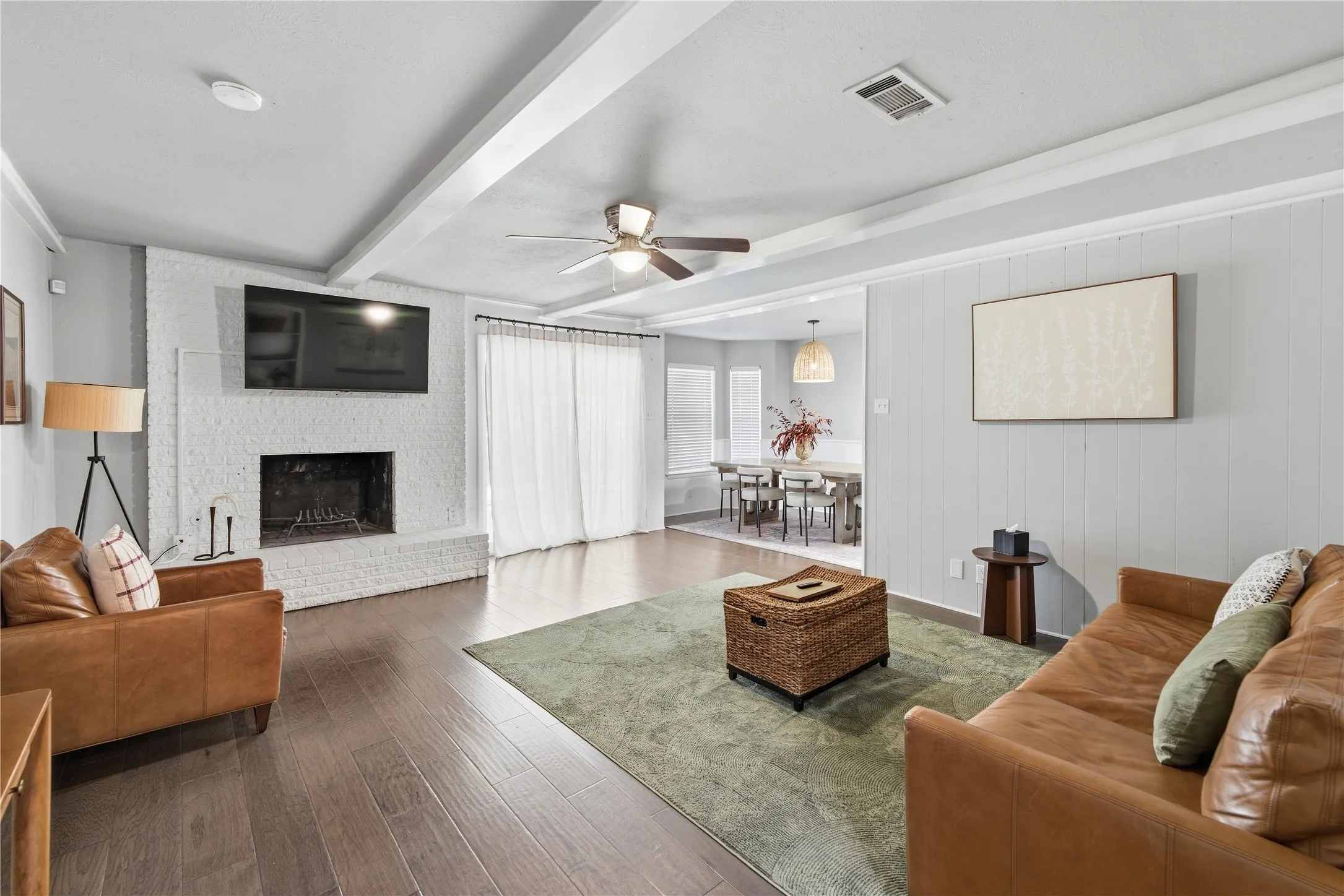 Spacious living area featuring wood-finish flooring, a white brick fireplace, and exposed ceiling beams