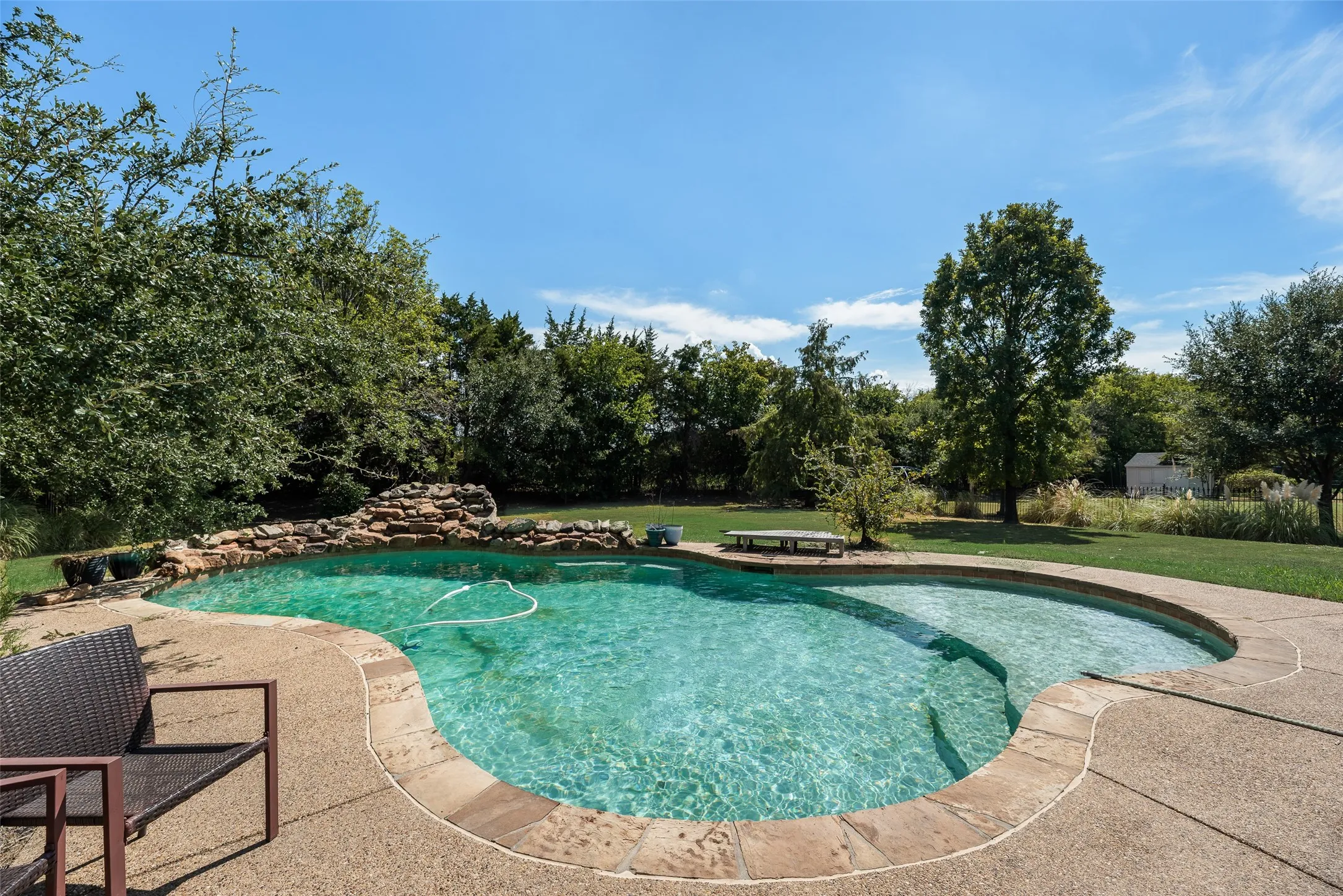 In-ground swimming pool with a rock waterfall feature, concrete pool deck, integrated spa area, and extensive surrounding greenery