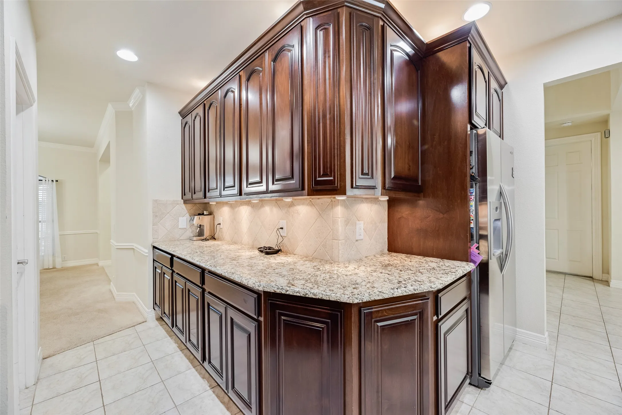 Dark wood-finish cabinetry with crown molding, light-colored speckled countertops, a patterned tile backsplash, and recessed under-cabinet lighting