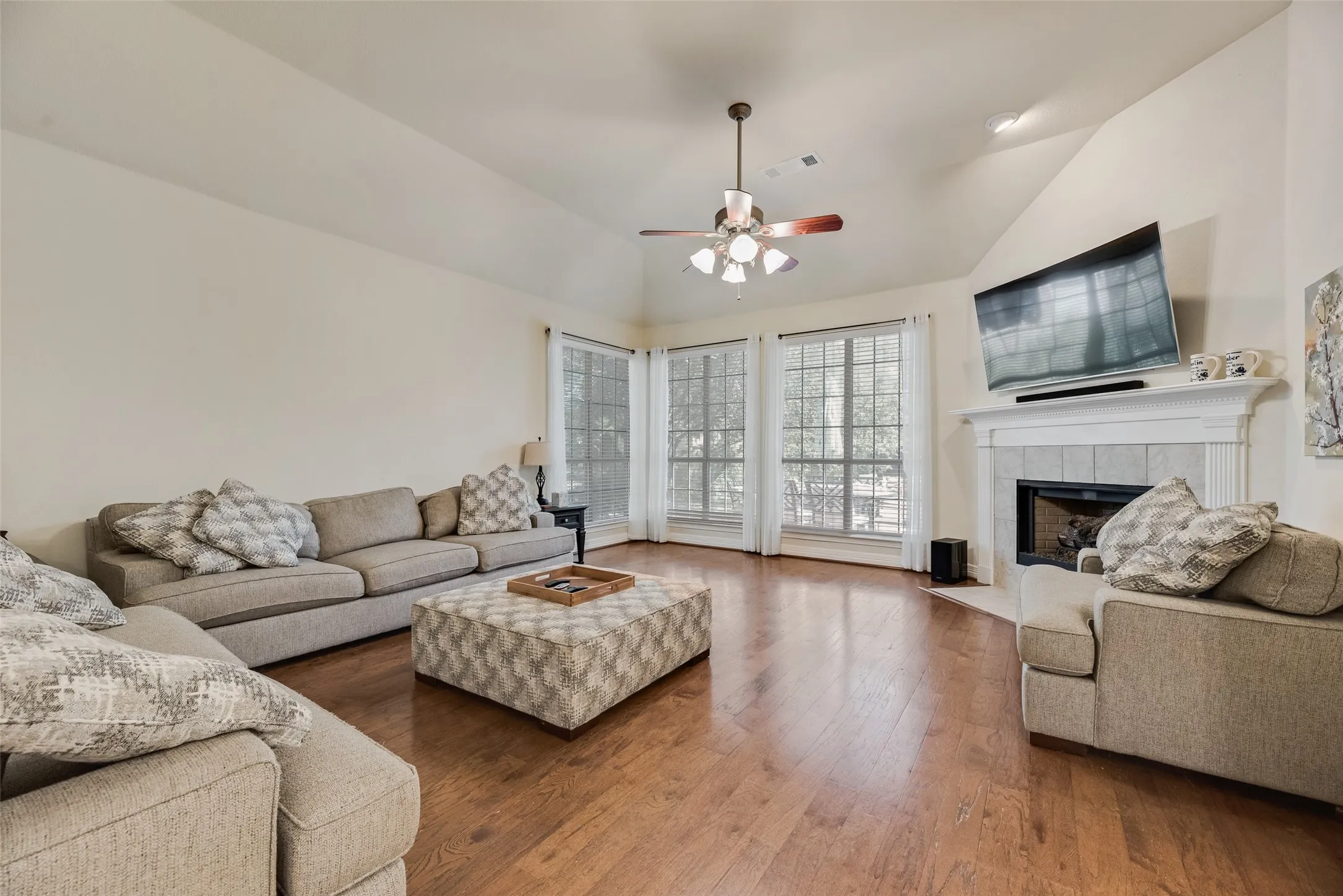 Vaulted ceiling with a ceiling fan light fixture, wood-finish flooring, multiple pane windows, and a tile fireplace with a mantel