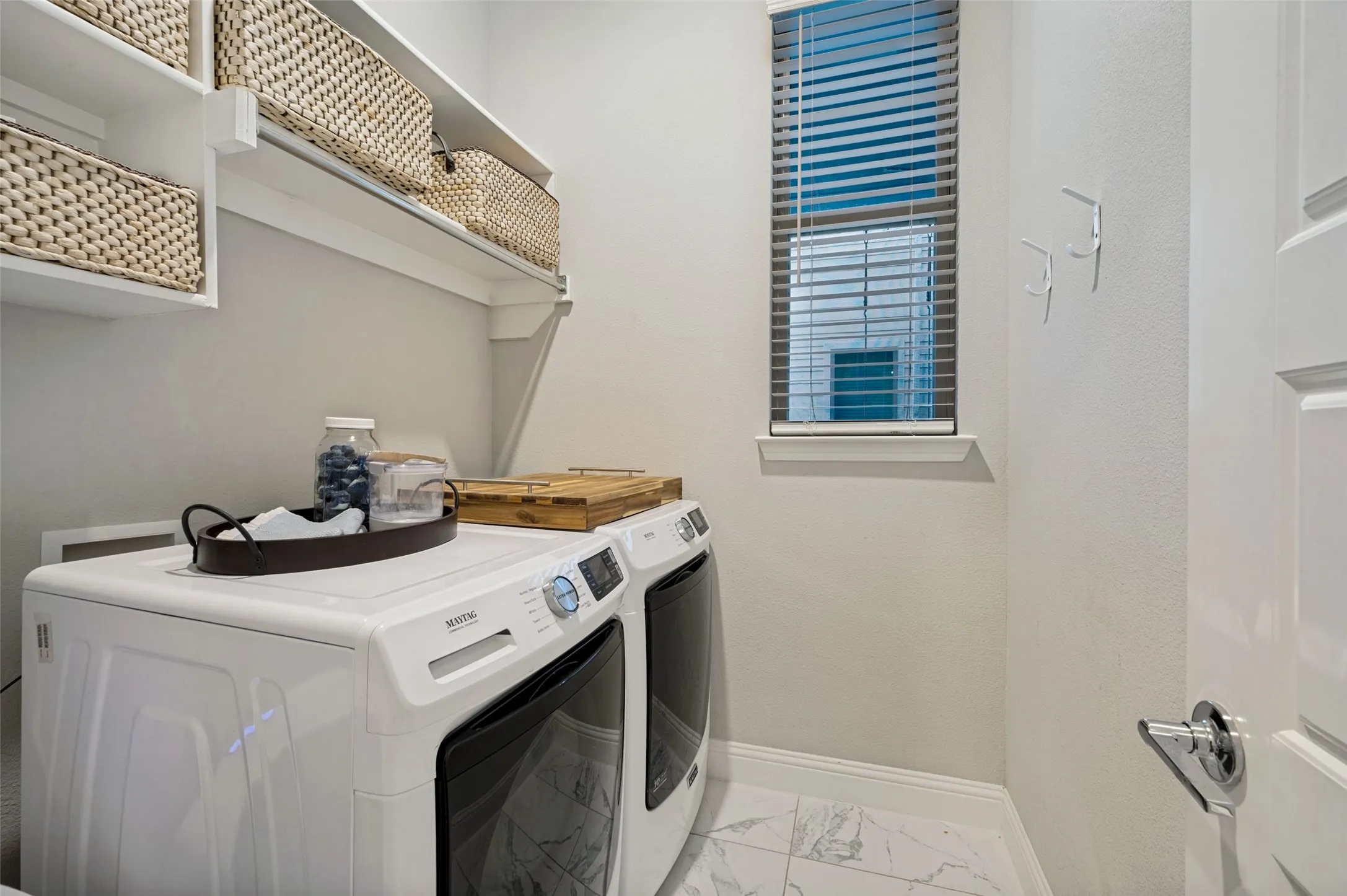 Laundry area featuring white shelving, a window with blinds, and marble-look tile flooring