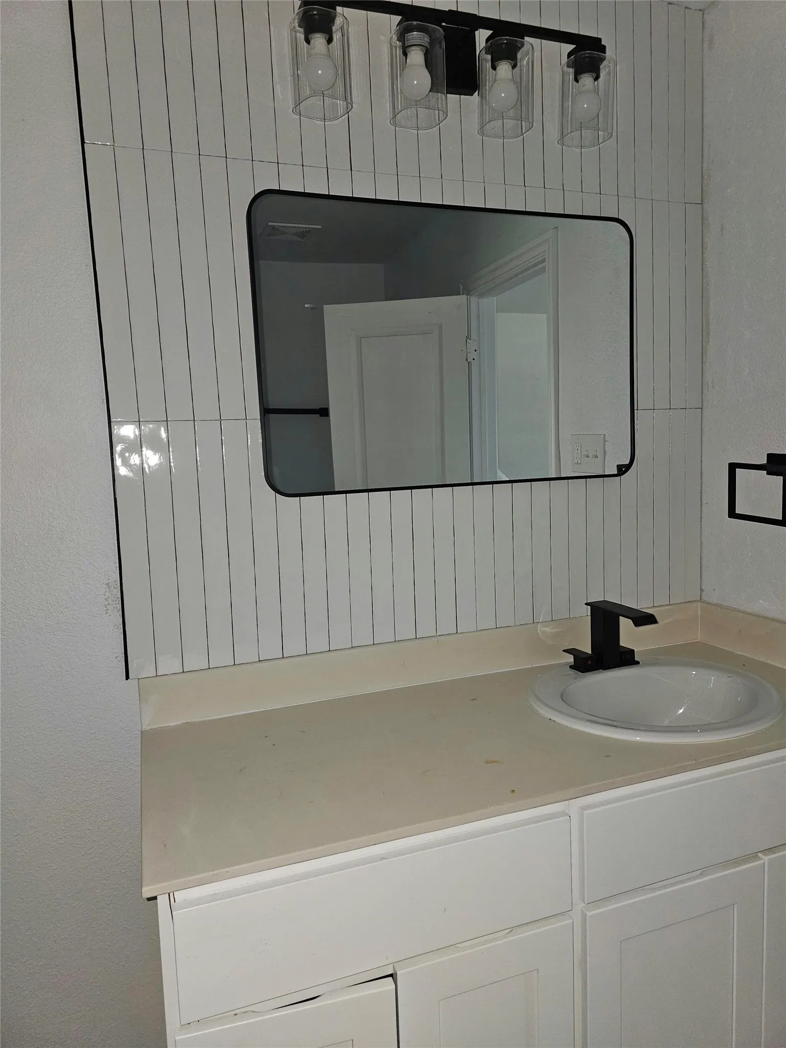 White subway tile backsplash, light-toned countertop, white under-sink cabinetry, black framed mirror, and a contemporary black four-light fixture