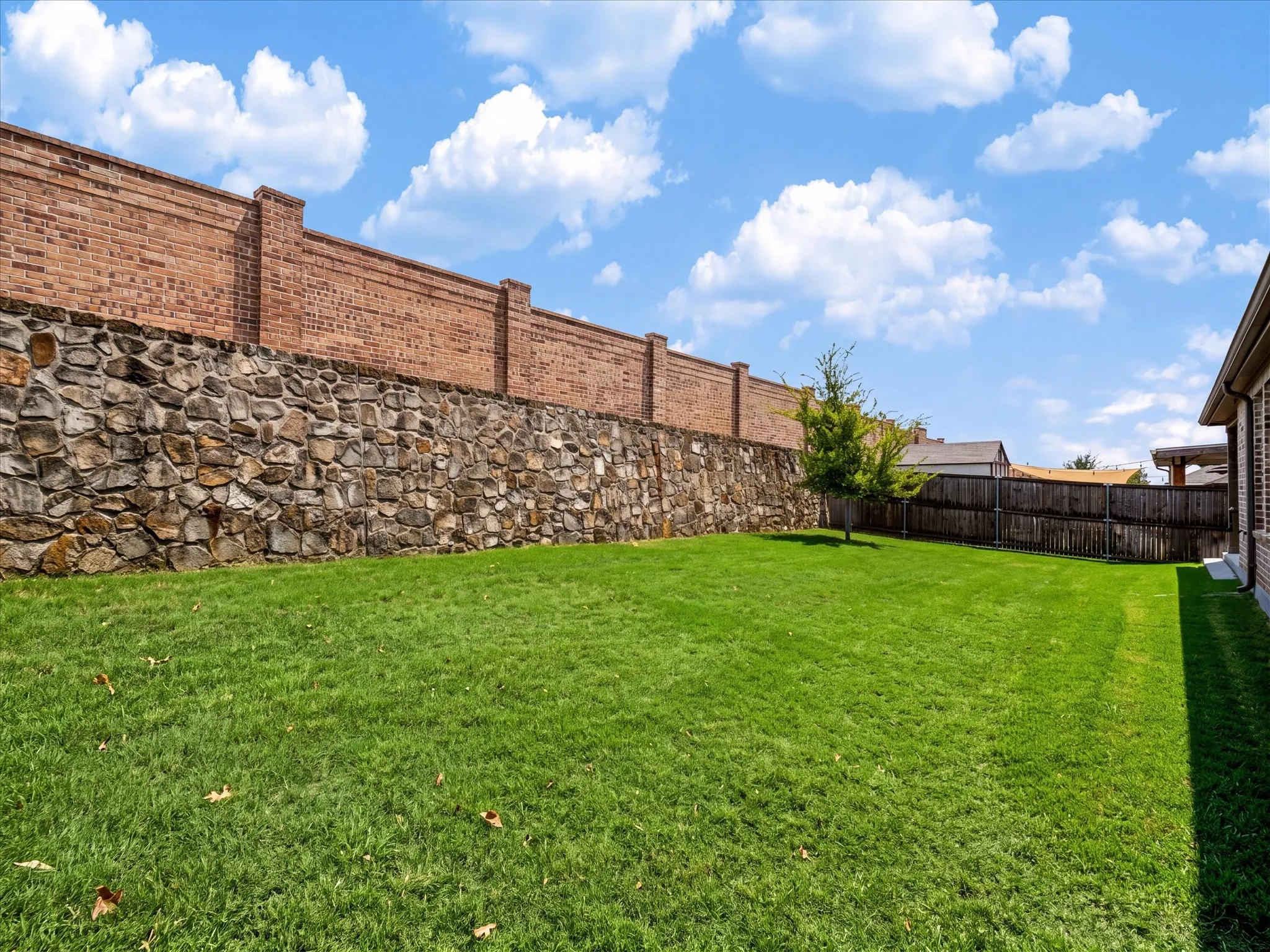 Expansive backyard featuring a manicured lawn with mature landscaping, bordered by a substantial brick and stone retaining wall that provides privacy and structure.  Digitally enhanced to show green grass.