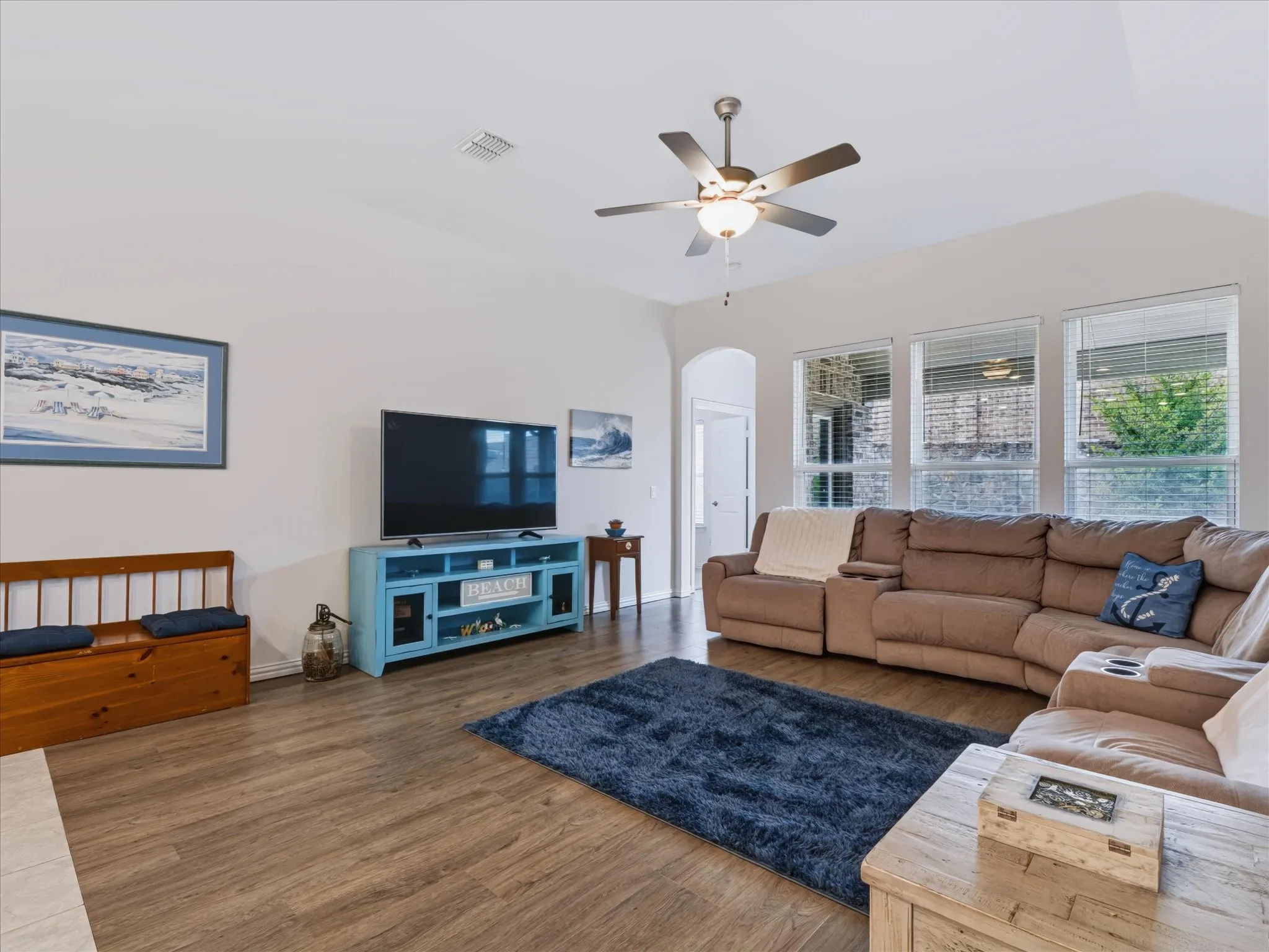 Wood-finish flooring, vaulted ceiling, ceiling fan with integrated lighting, multiple windows, and an arched doorway