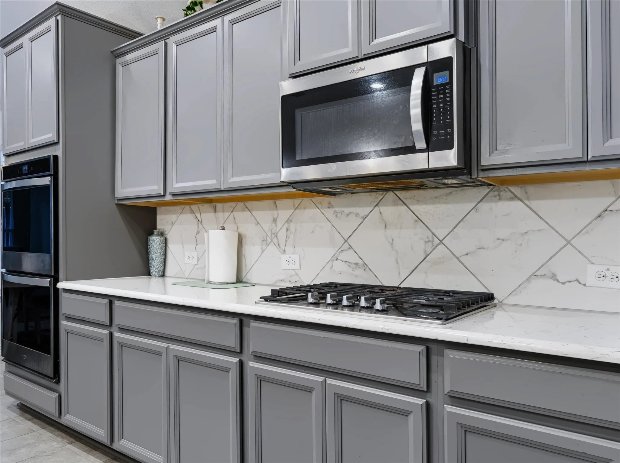 Gray shaker cabinetry, built-in double ovens, stainless steel microwave, gas cooktop, and white countertops with a patterned backsplash