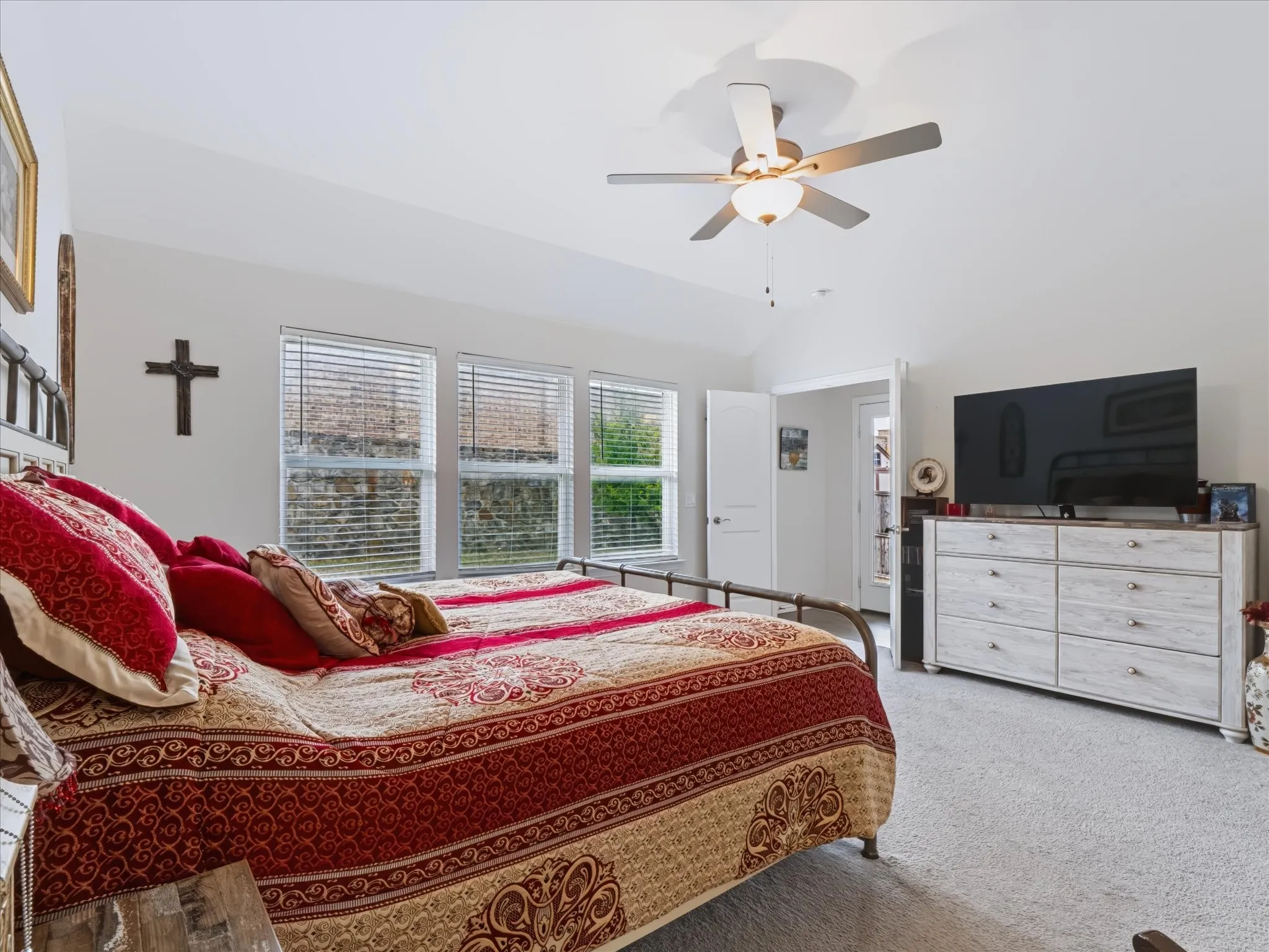 Vaulted ceiling, ceiling fan, three windows with blinds, light-toned carpeting, and a white interior door