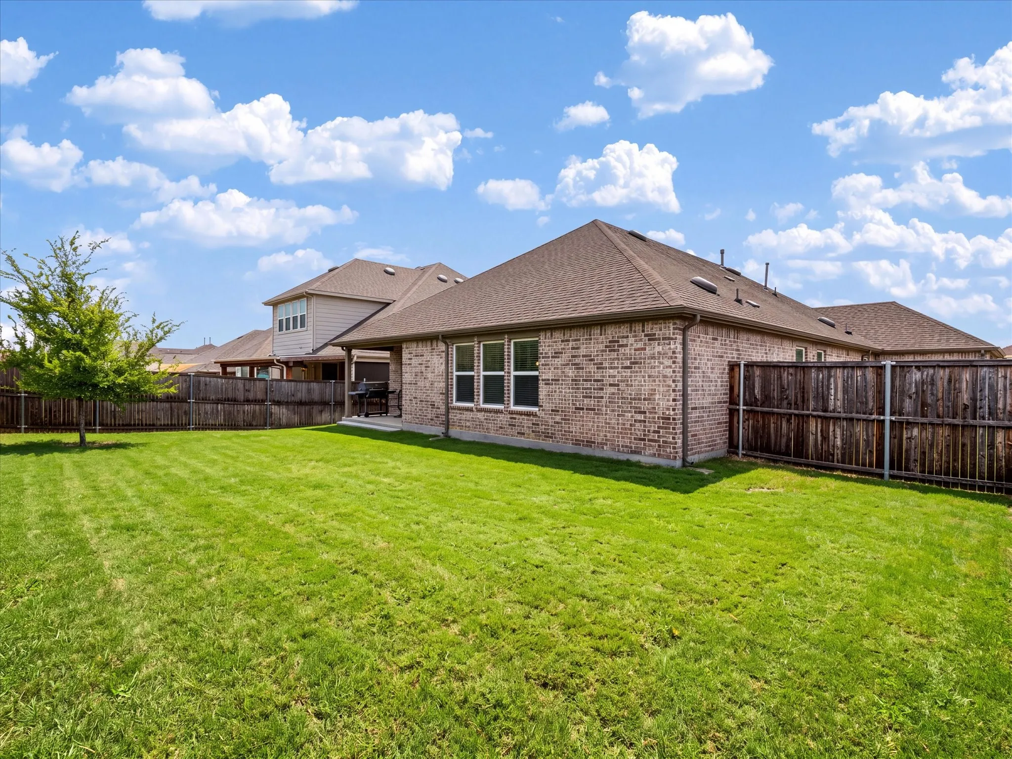 Expansive lawn with a mature tree, brick exterior, brown shingle roof, and a covered patio.  Digitally enhanced to show green grass.