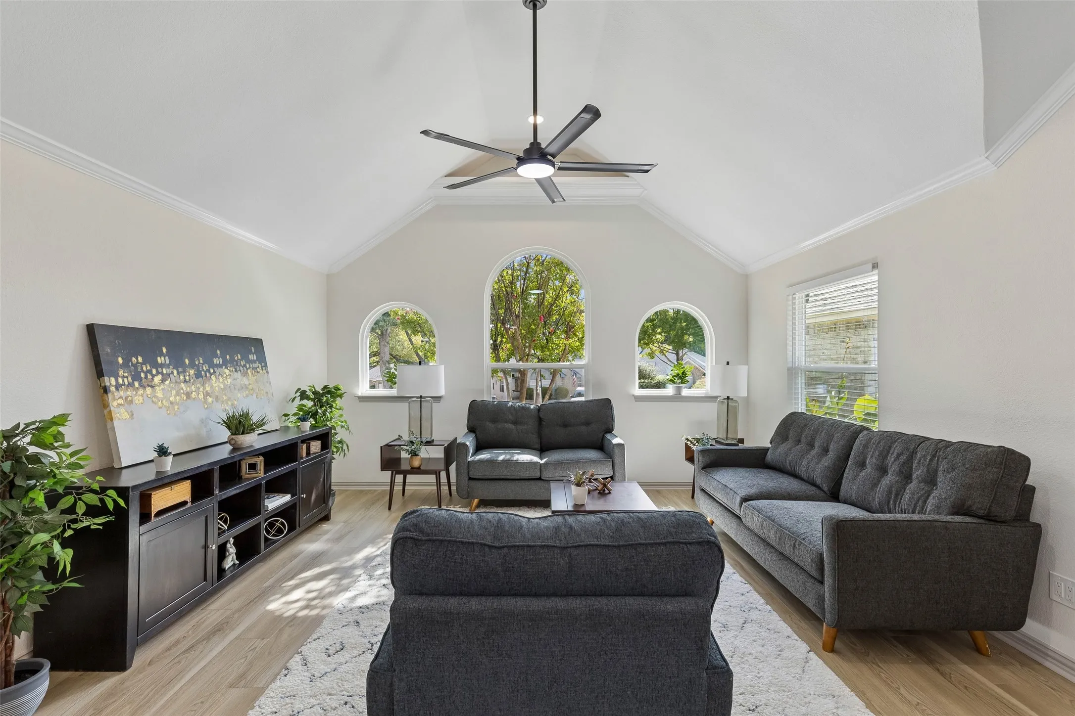 Vaulted ceiling with a contemporary ceiling fan, three arched windows, crown molding, wood-finish flooring, and a built-in media console