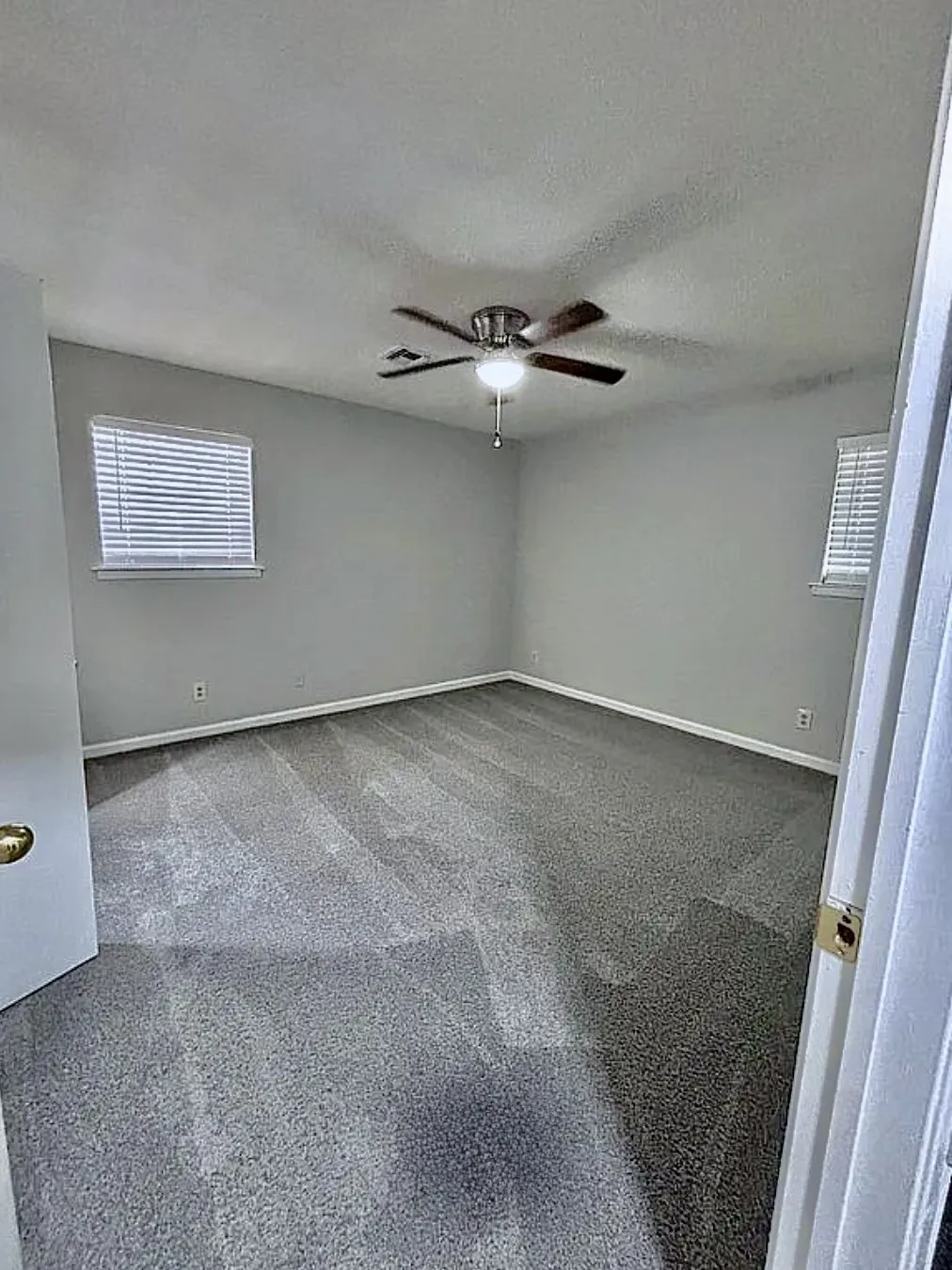 Neutral wall paint, ceiling fan with integrated lighting, two windows with blinds, and gray carpeting