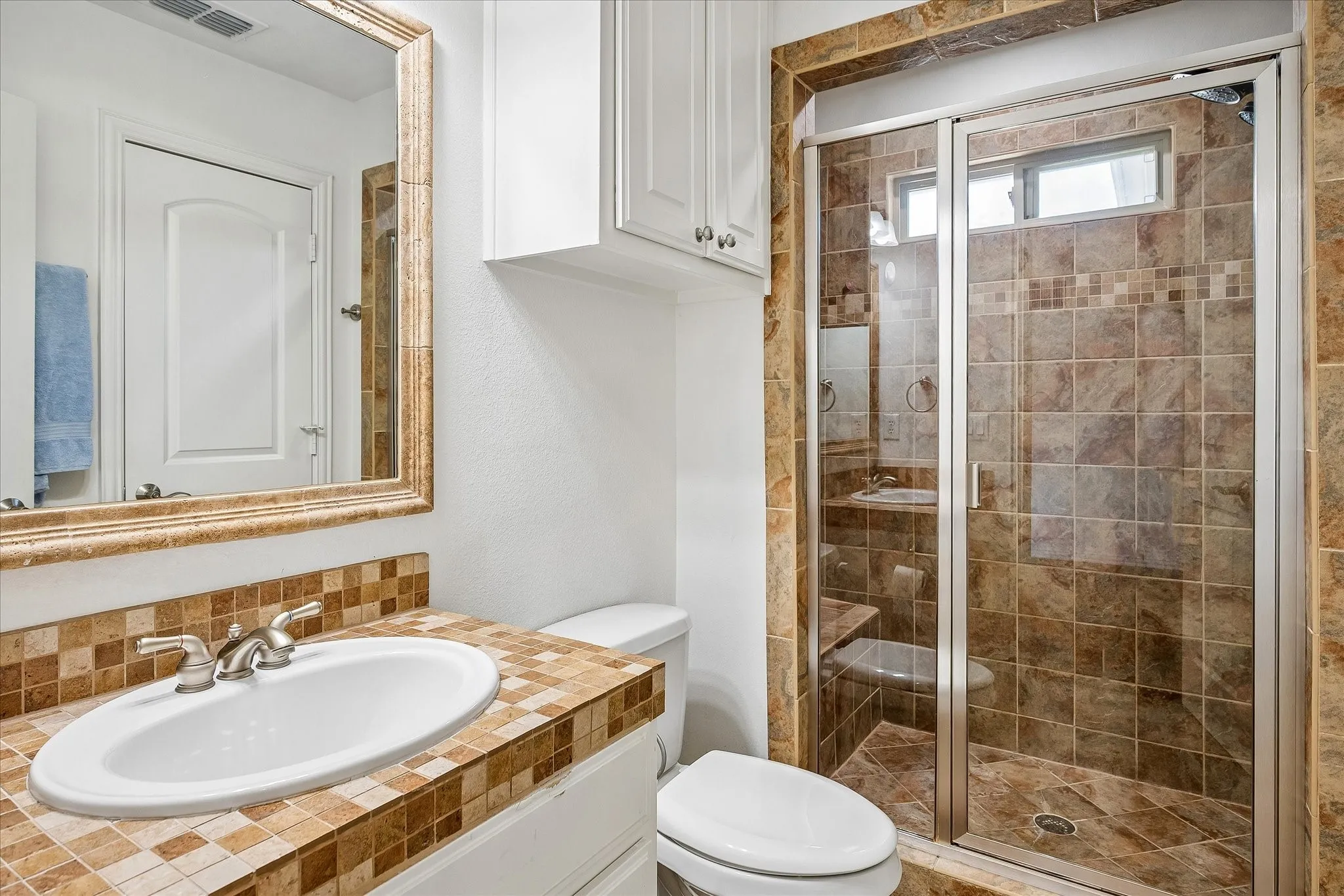 Tiled vanity countertop with integrated sink, framed mirror, white cabinetry, and a walk-in shower with tiled surround and glass enclosure