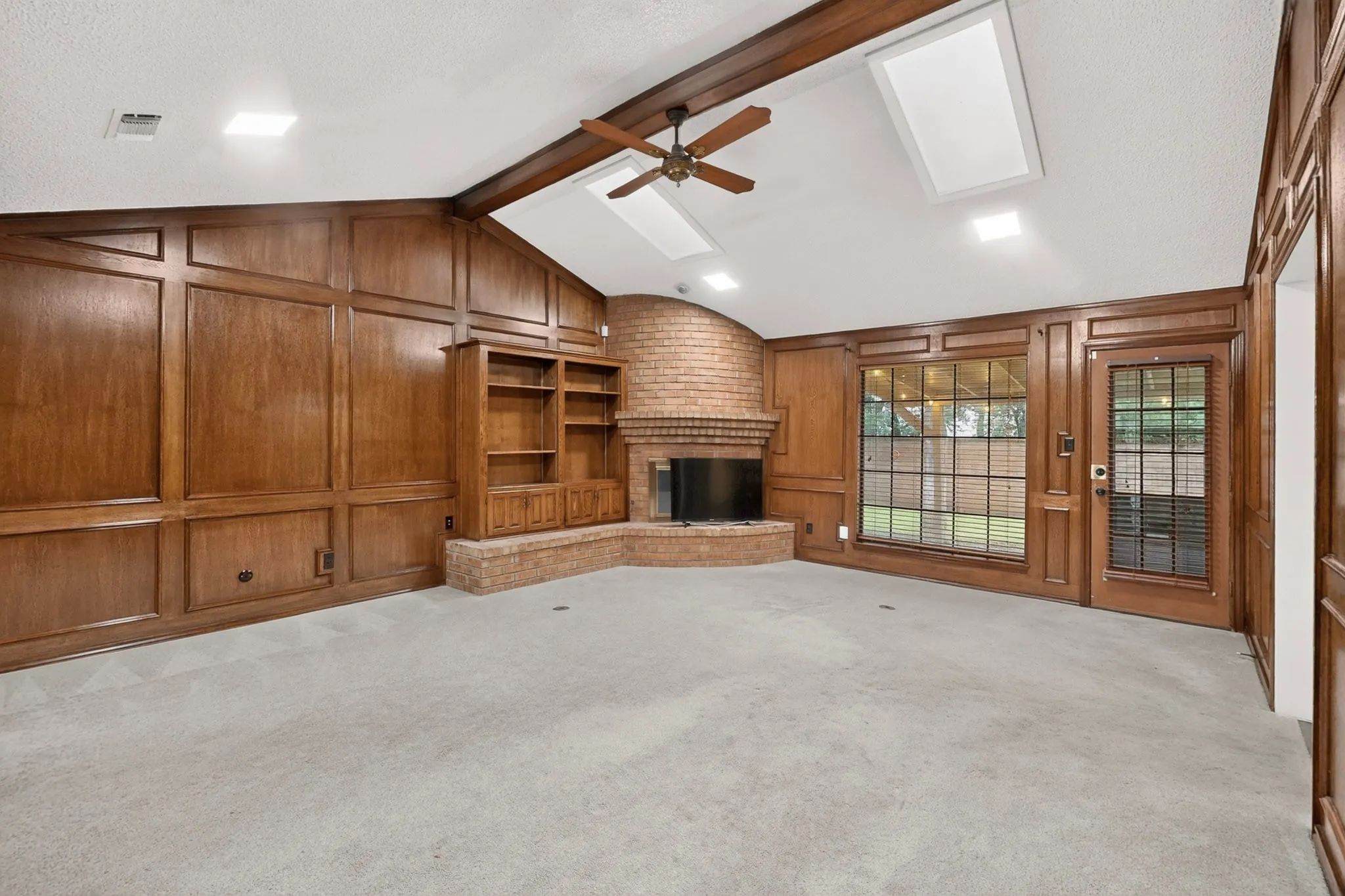 Vaulted ceilings with exposed wood beams, brick fireplace, built-in shelving, wood-paneled walls, and exterior access via a glass-paneled door