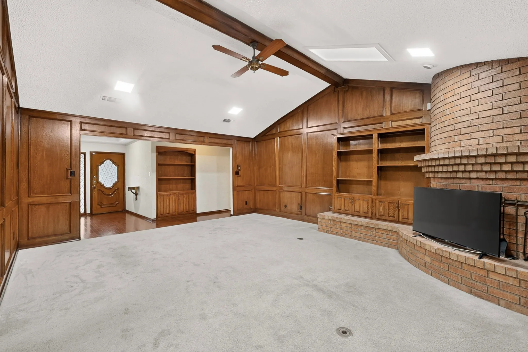 Vaulted ceiling with exposed wood beams, wood-paneled walls with built-in cabinetry, brick fireplace, and wood-finish entry door with decorative glass