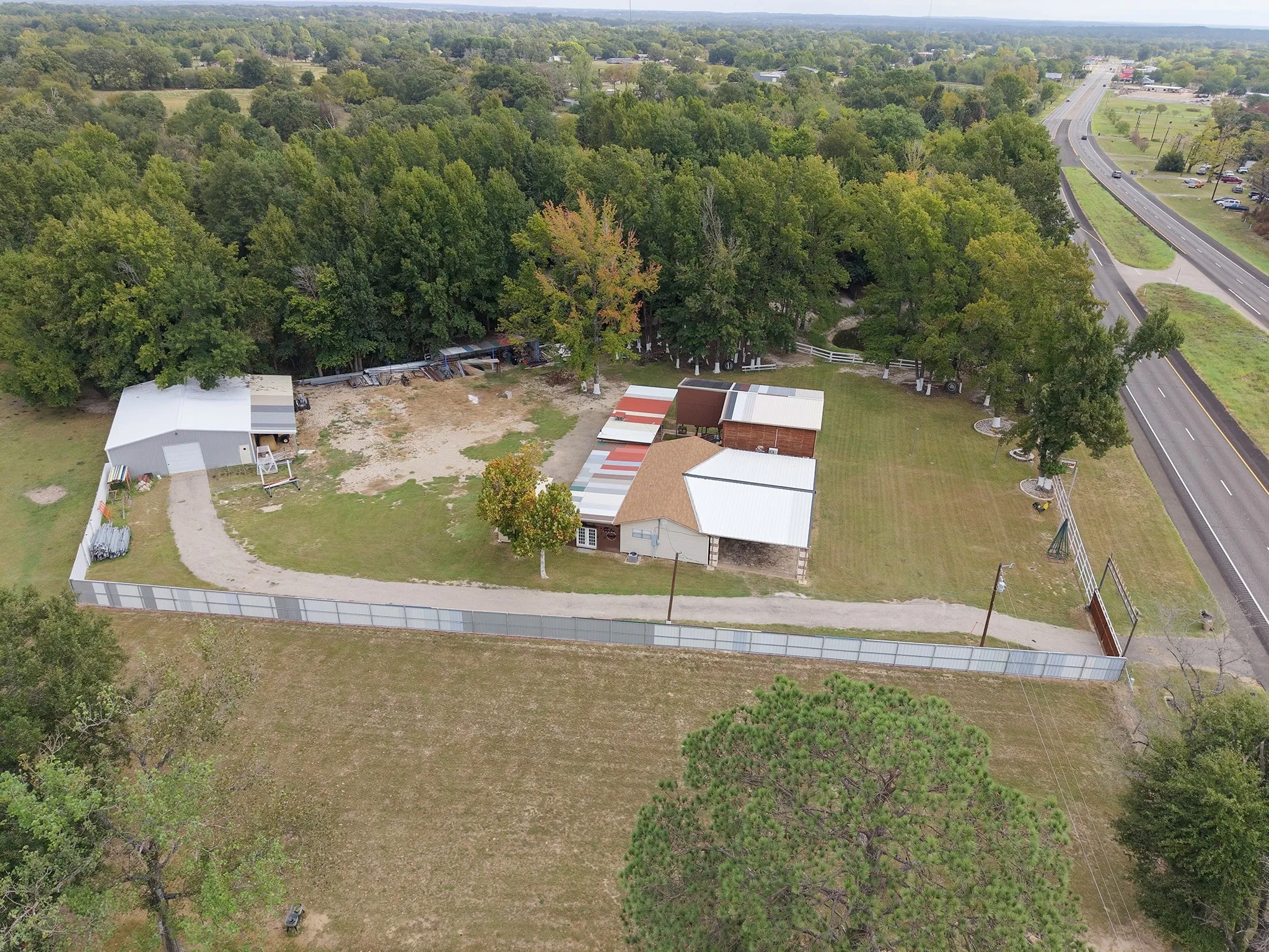 Gated property featuring a main structure with a light-colored roof, a separate metal-sided outbuilding, a gravel driveway, and mature perimeter trees
