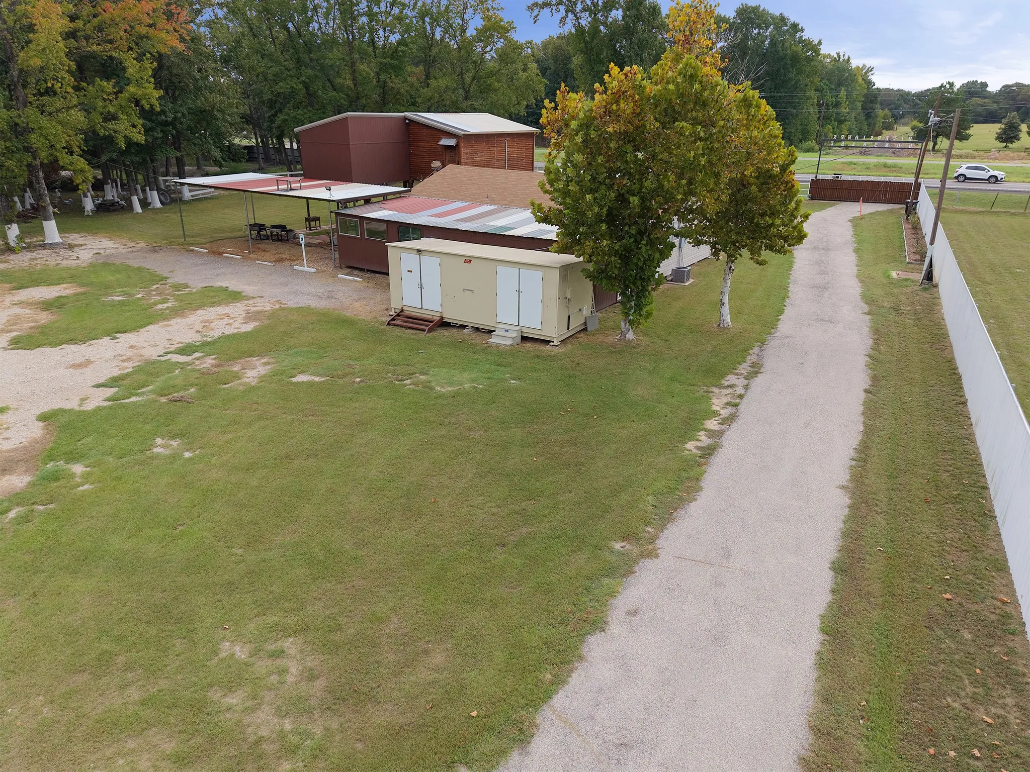 Expansive grounds with a gravel driveway, mature trees, a perimeter fence, and multiple outbuildings