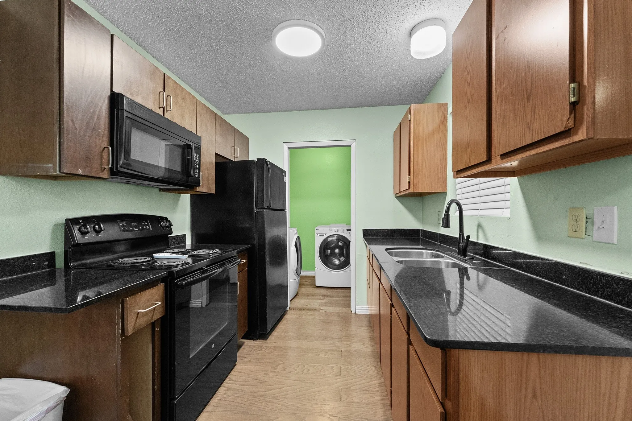 Galley kitchen featuring wood cabinetry, black appliances, dark countertops, and wood-finish flooring