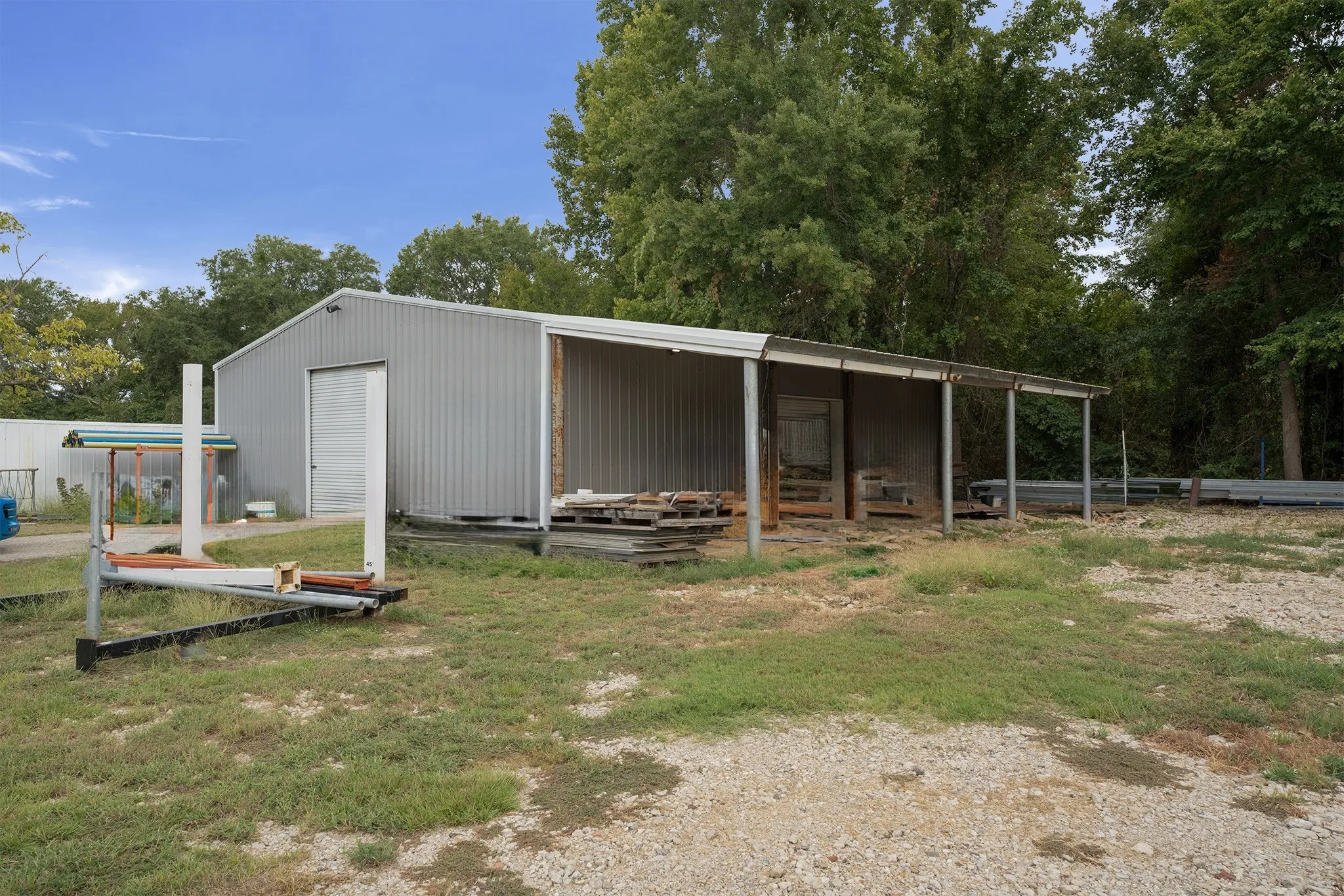 Metal-sided building featuring a large roll-up door, an attached open-sided covered area, and a gravel surface