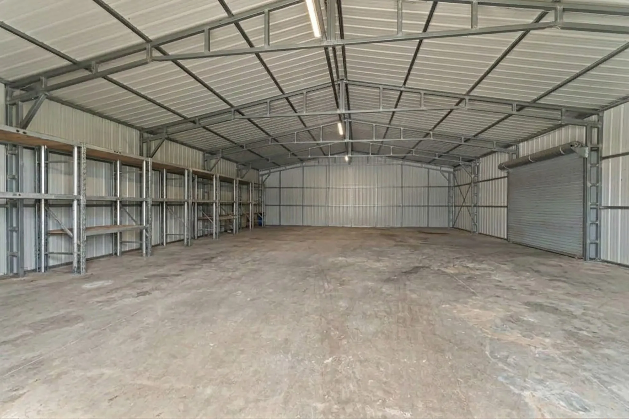 Expansive metal structure featuring a concrete floor, built-in shelving, corrugated metal walls, a roll-up door, and exposed metal trusses