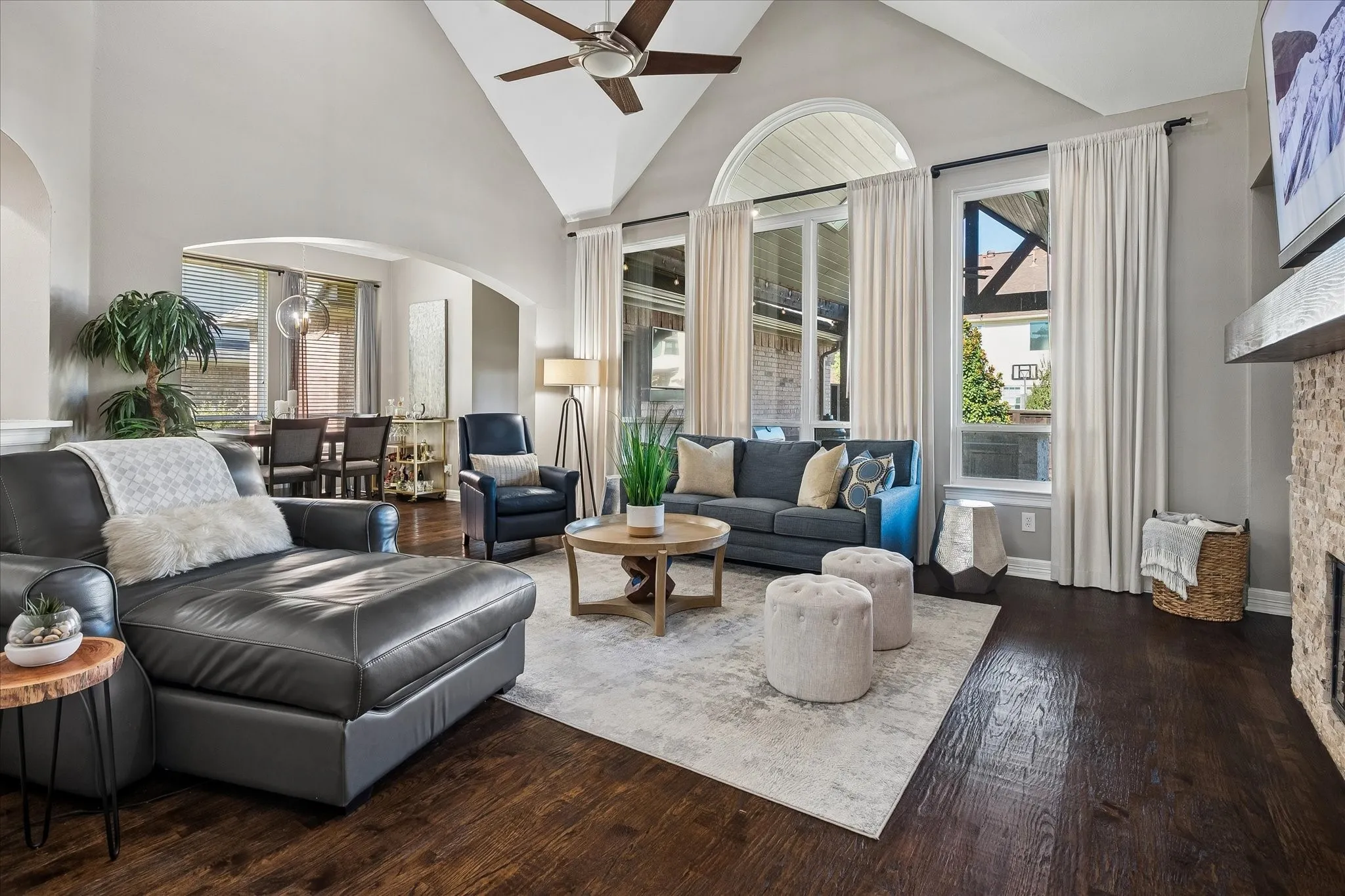 Vaulted ceiling with a ceiling fan, dark wood-finish flooring, stone-clad fireplace with a wood mantel, and tall windows
