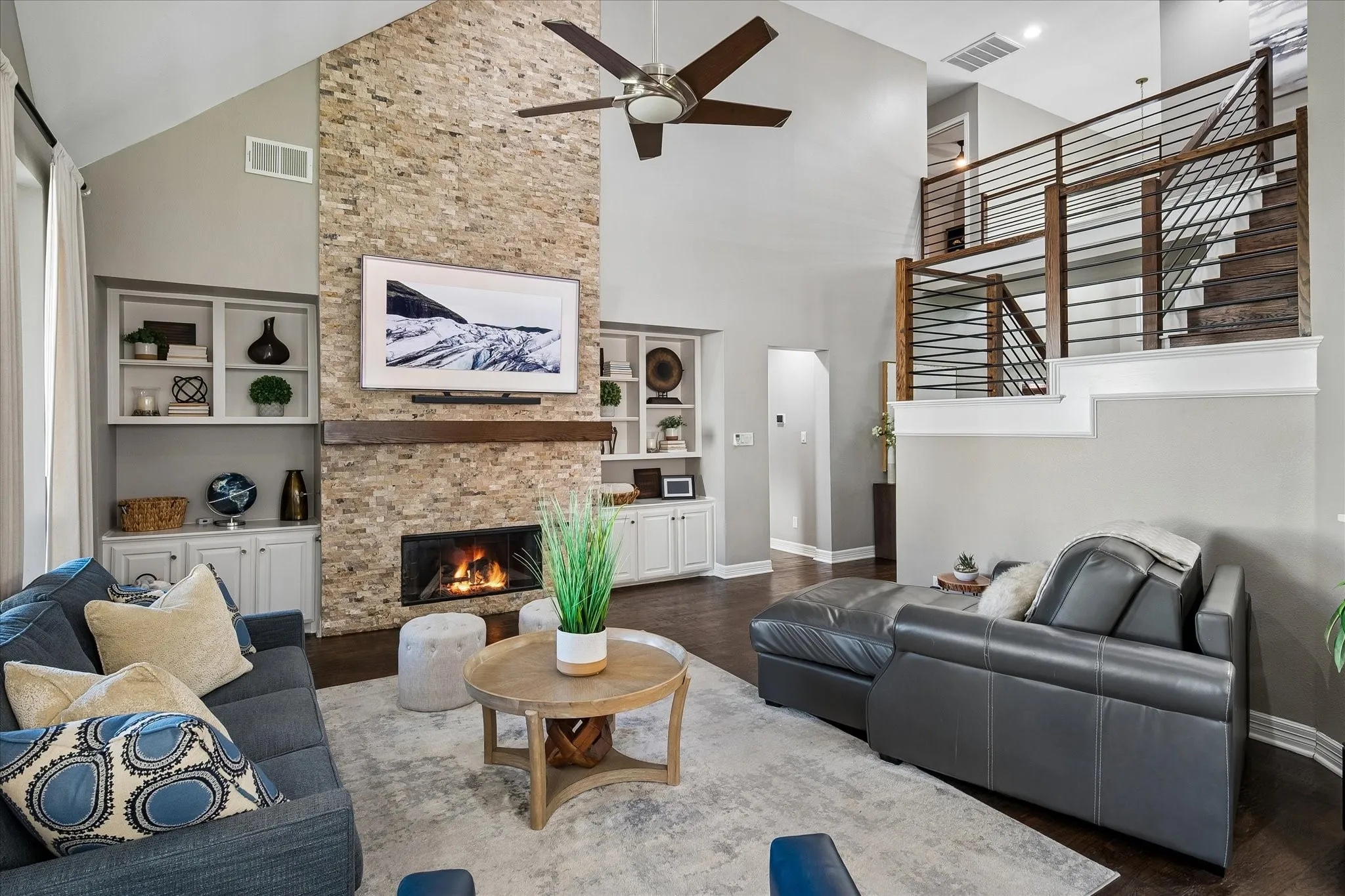 Vaulted ceiling with a ceiling fan, stone-clad fireplace with wood mantel, built-in shelving, wood-finish flooring, and a modern staircase with wood and metal railing