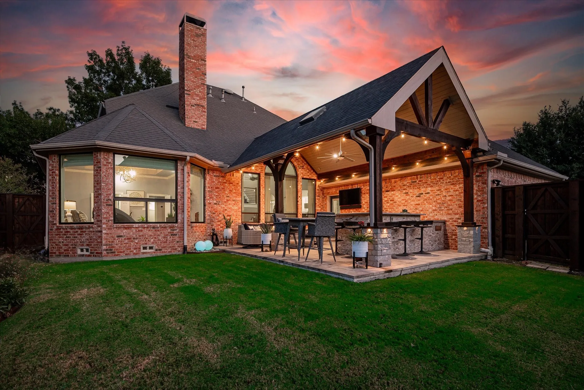 Brick exterior with a prominent chimney, large bay windows, and a covered patio featuring exposed wood beams, a built-in outdoor kitchen, and string lighting